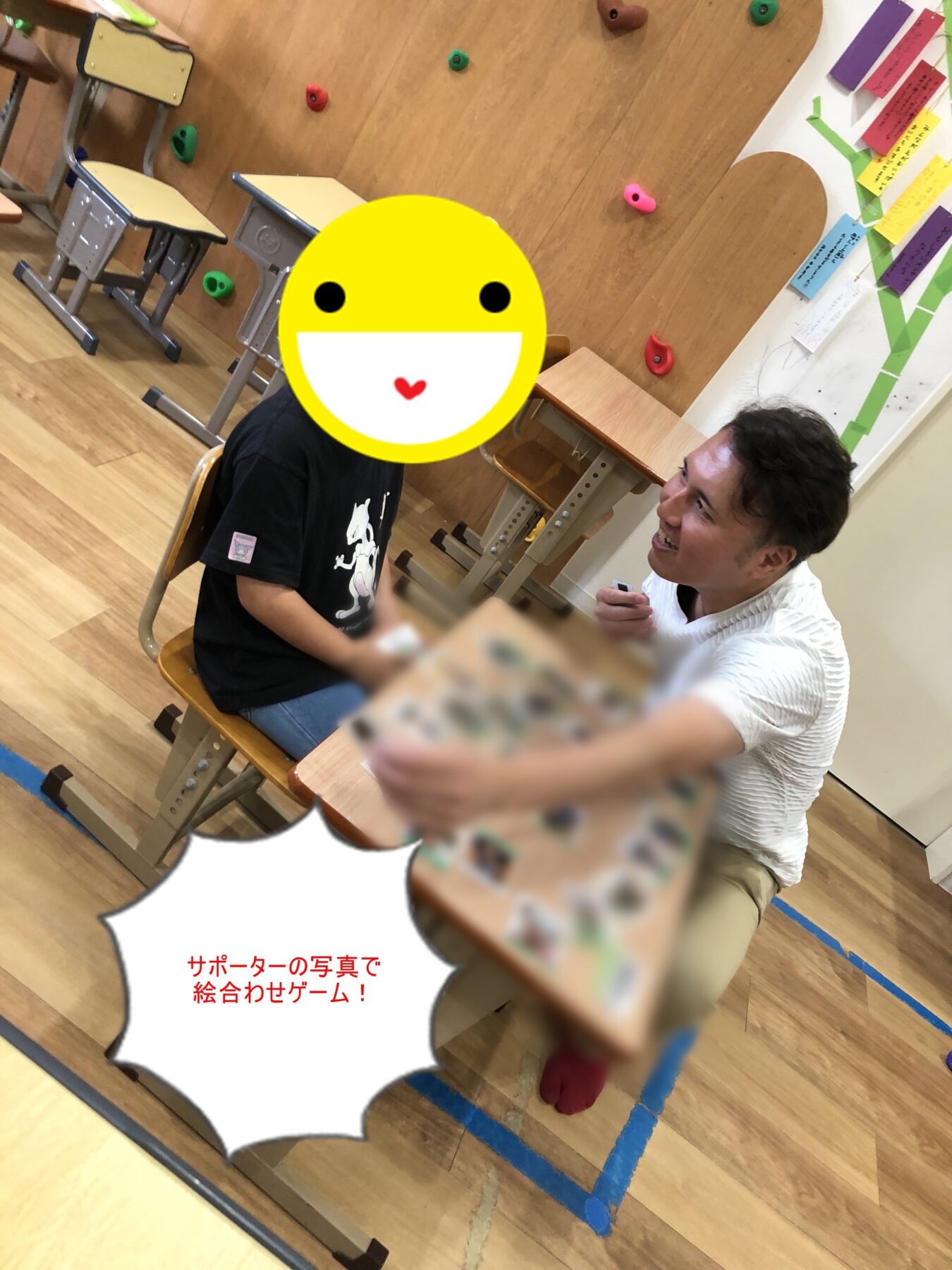 Teacher or volunteer smiles and hands a card to a student during a classroom activity; desks and a climbing wall with colorful holds visible in the background.