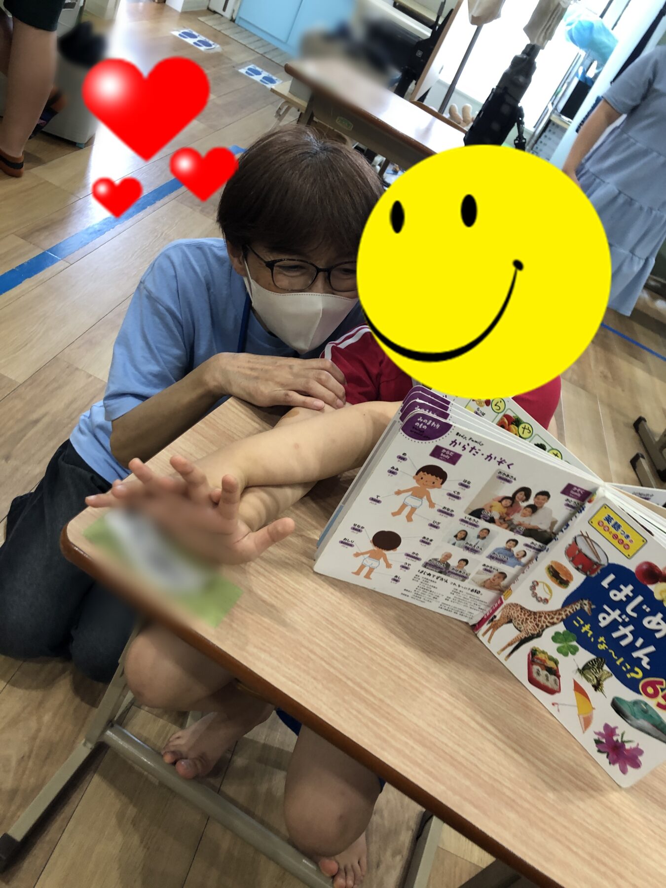 Adult in a blue shirt wearing a mask helps a child at a table with an open illustrated book in a classroom or clinic setting; large yellow smiley face and red hearts obscure the faces, focusing on interaction and activity.
