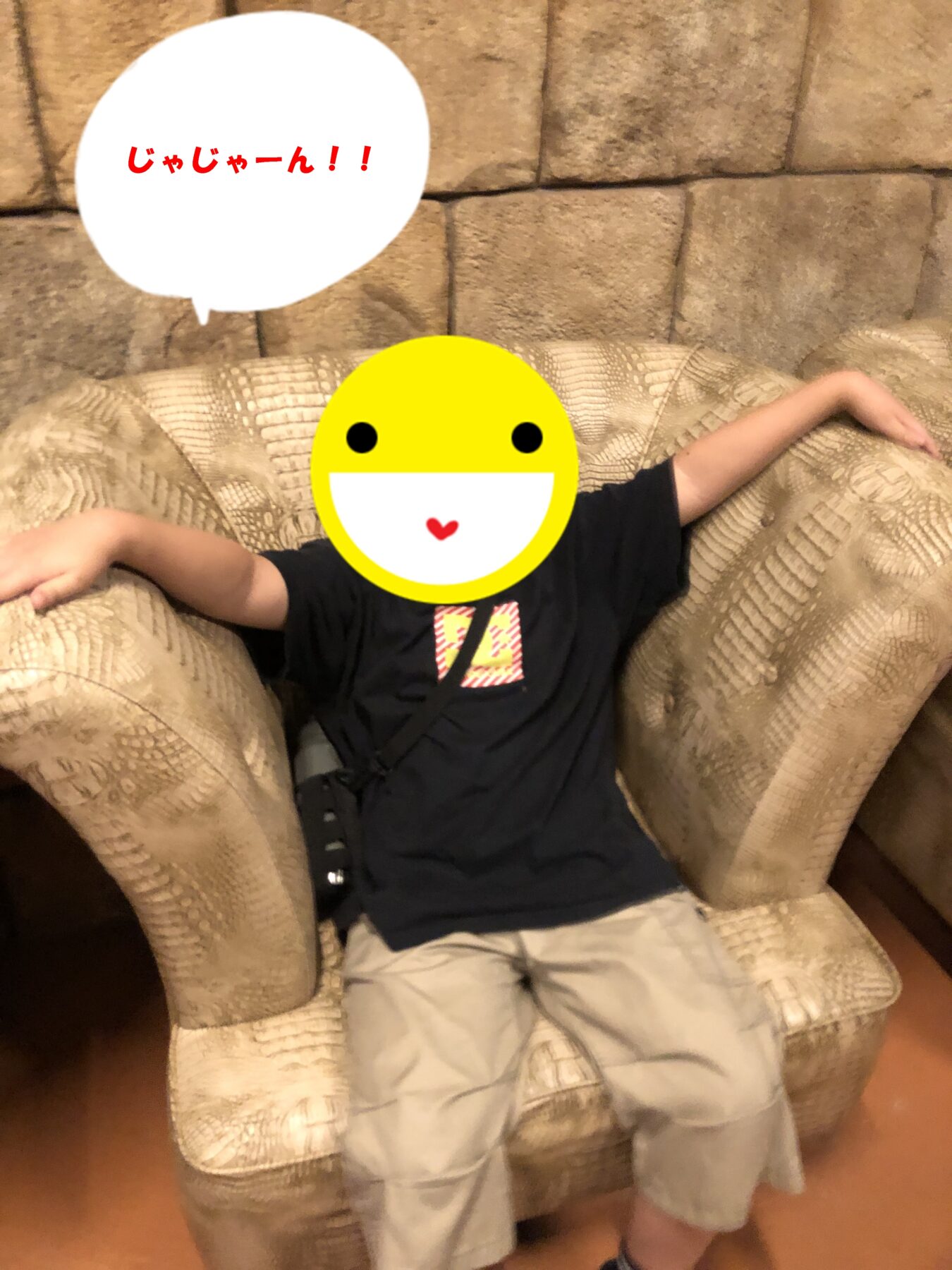 Child sitting on a beige patterned sofa with face covered by a yellow smiley emoji; speech bubble shows Japanese text じゃじゃーん!!