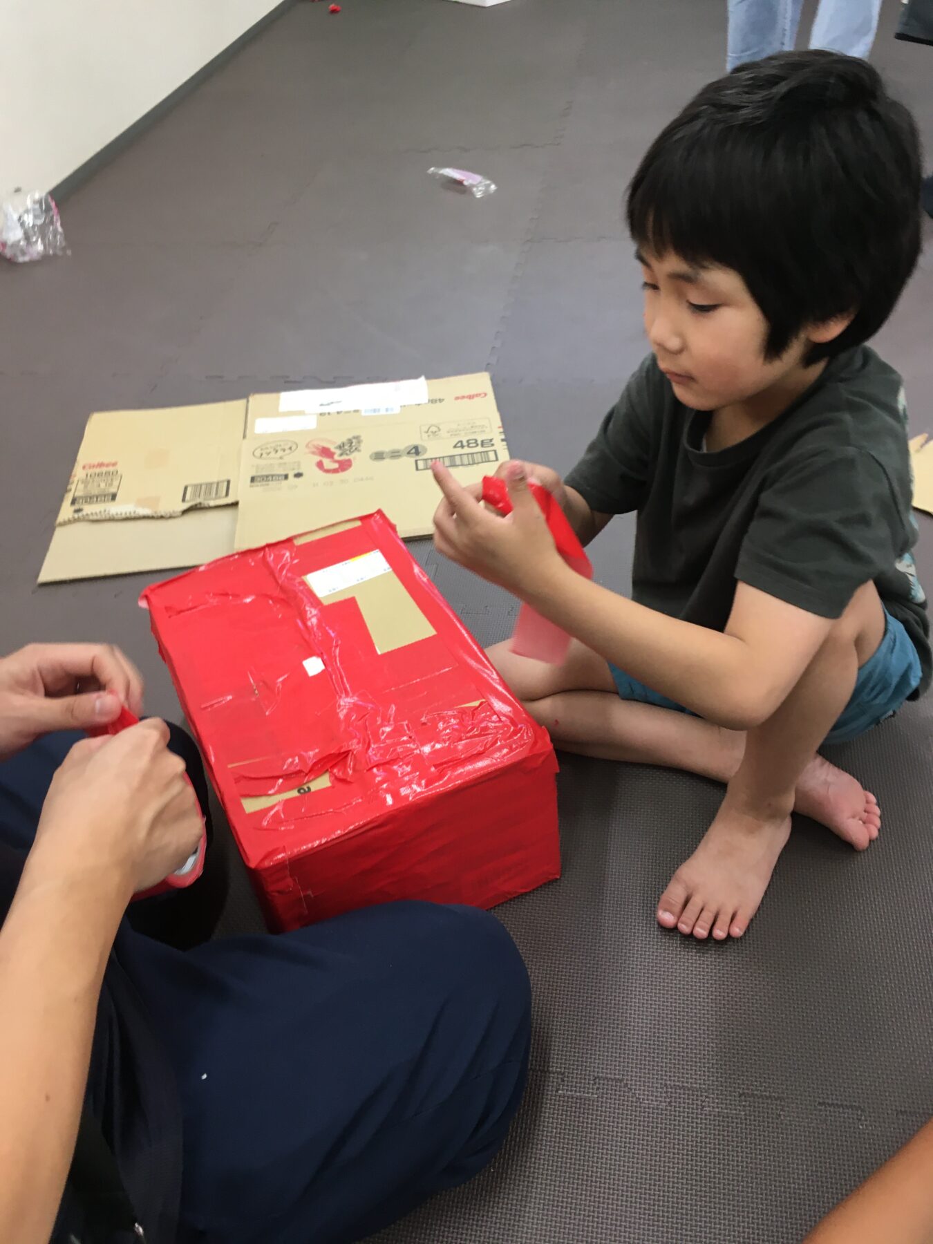 Young child with dark hair sits on a gray gym mat, unwrapping a bright red gift while another person helps nearby.