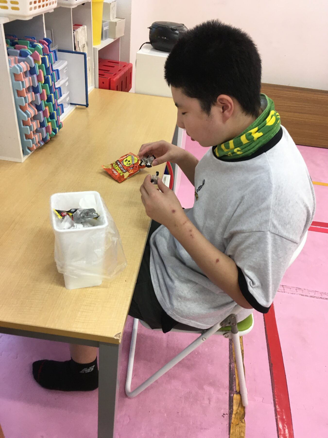 Person seated at a light wood table in a colorful classroom, opening a red snack bag with a small plastic bag nearby; shelves with colorful organizers in the background.