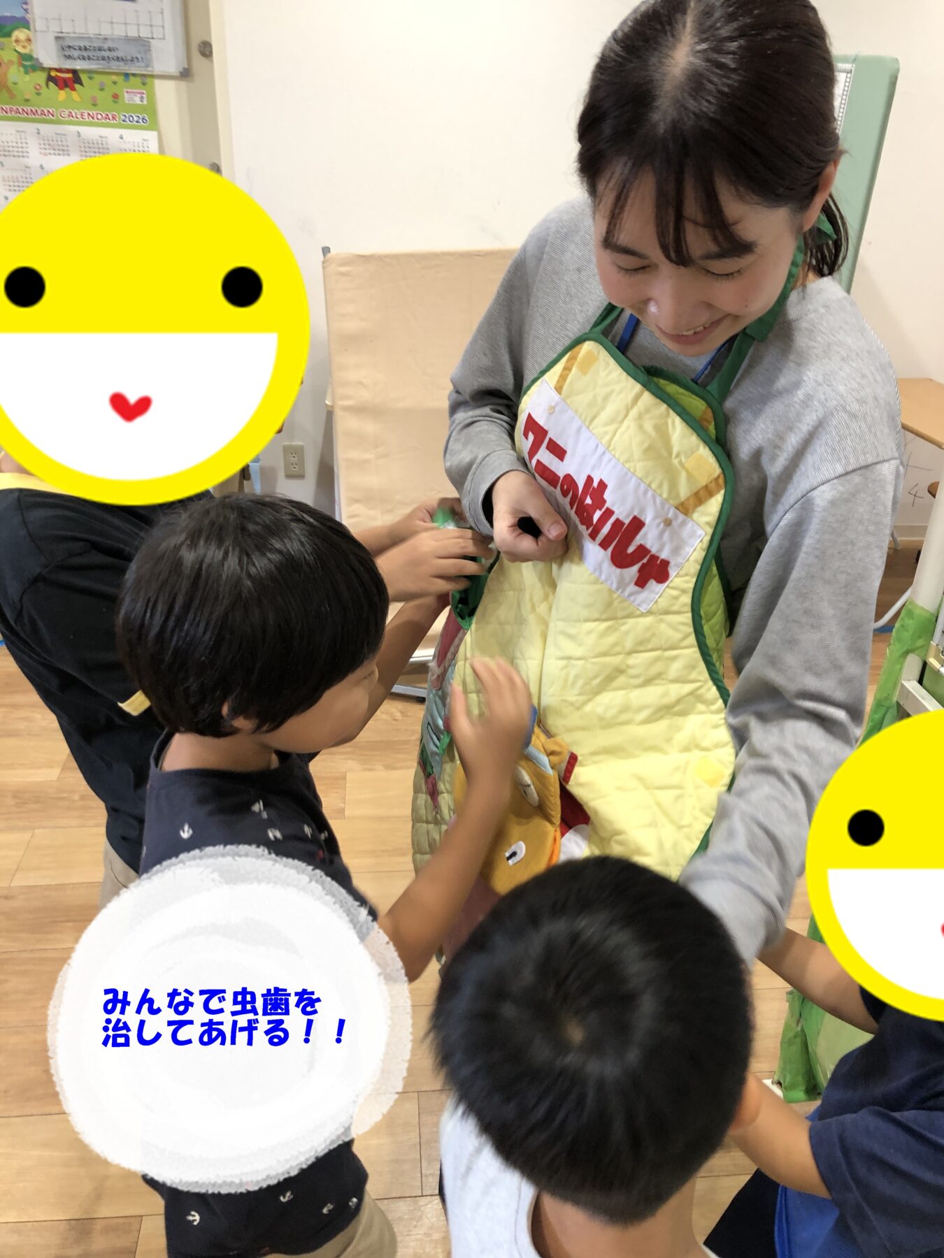 Children gather around a smiling instructor in a yellow quilted apron as they participate in a dental care activity in a classroom. A white circular sign with blue Japanese text sits in the foreground: 'みんなで虫歯を治してあげる!!'