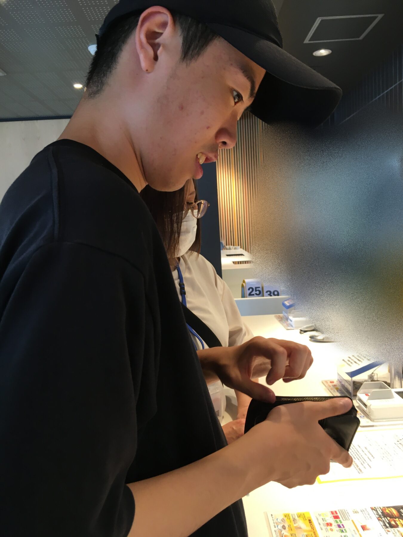 Side profile of a person in a black cap looking at a smartphone at a brightly lit counter, indoors.