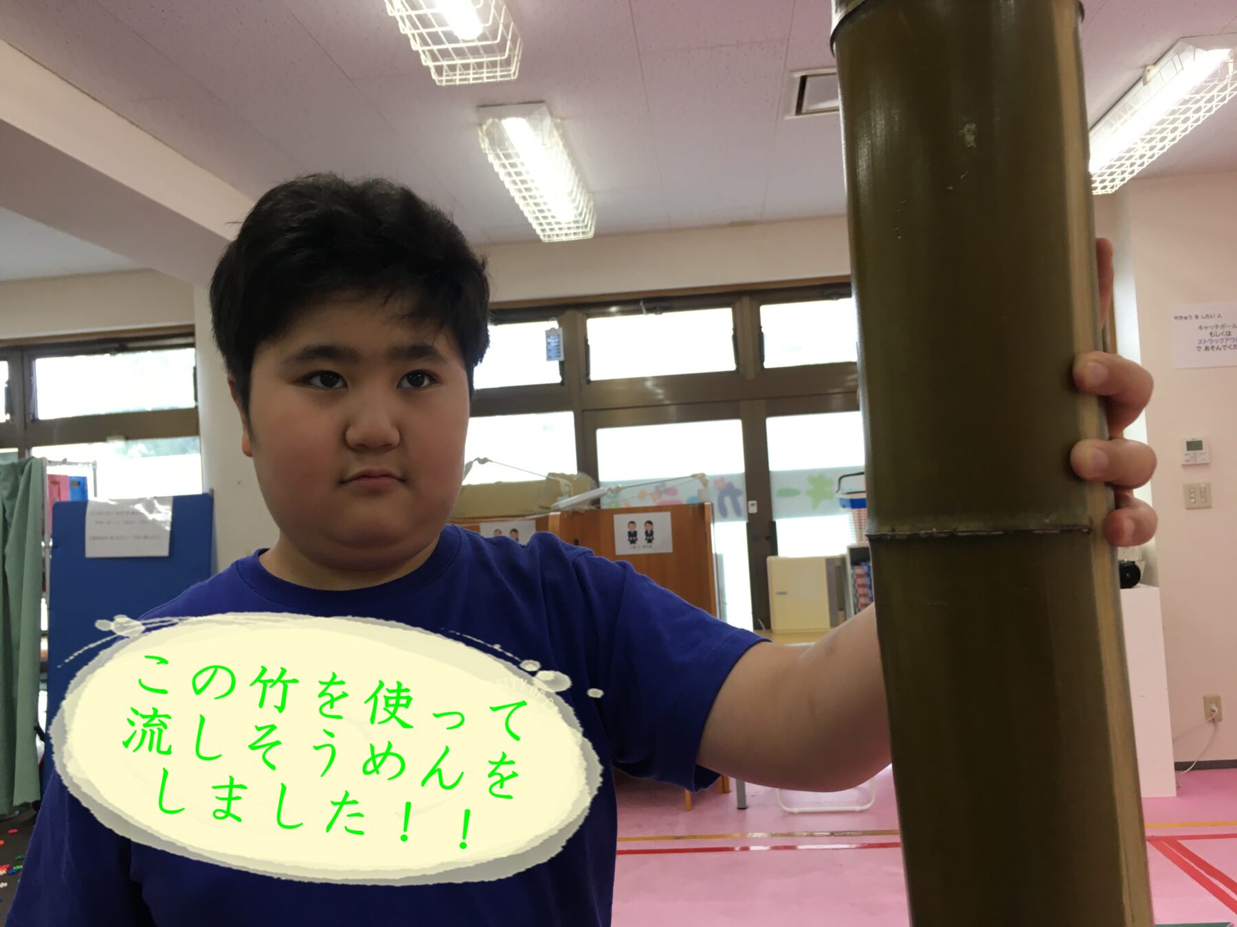 Person in a blue shirt holds a large bamboo pole indoors, with a speech-bubble message in Japanese: この竹を使って流しそうめんをしました！！
