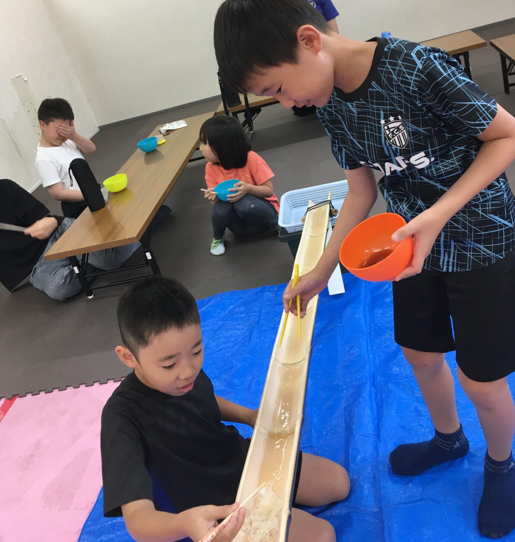 Children gathered around a wooden water chute; a boy in black sits on a pink mat watching the flowing water, with a blue bowl nearby.