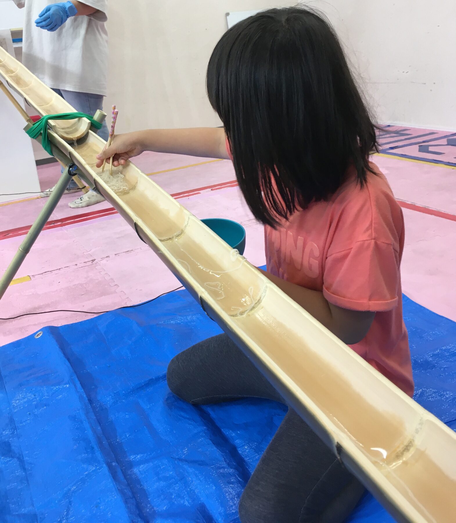Child in a pink shirt kneels beside a light bamboo water slide, touching the running water with sticks while a person in a white coat watches.