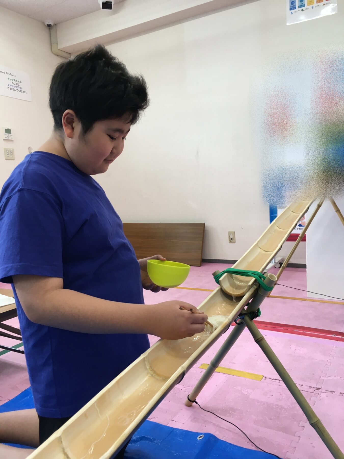 Young boy in a blue shirt stands beside a long wooden water trough, pouring water from a yellow bowl into it in an indoor play area.