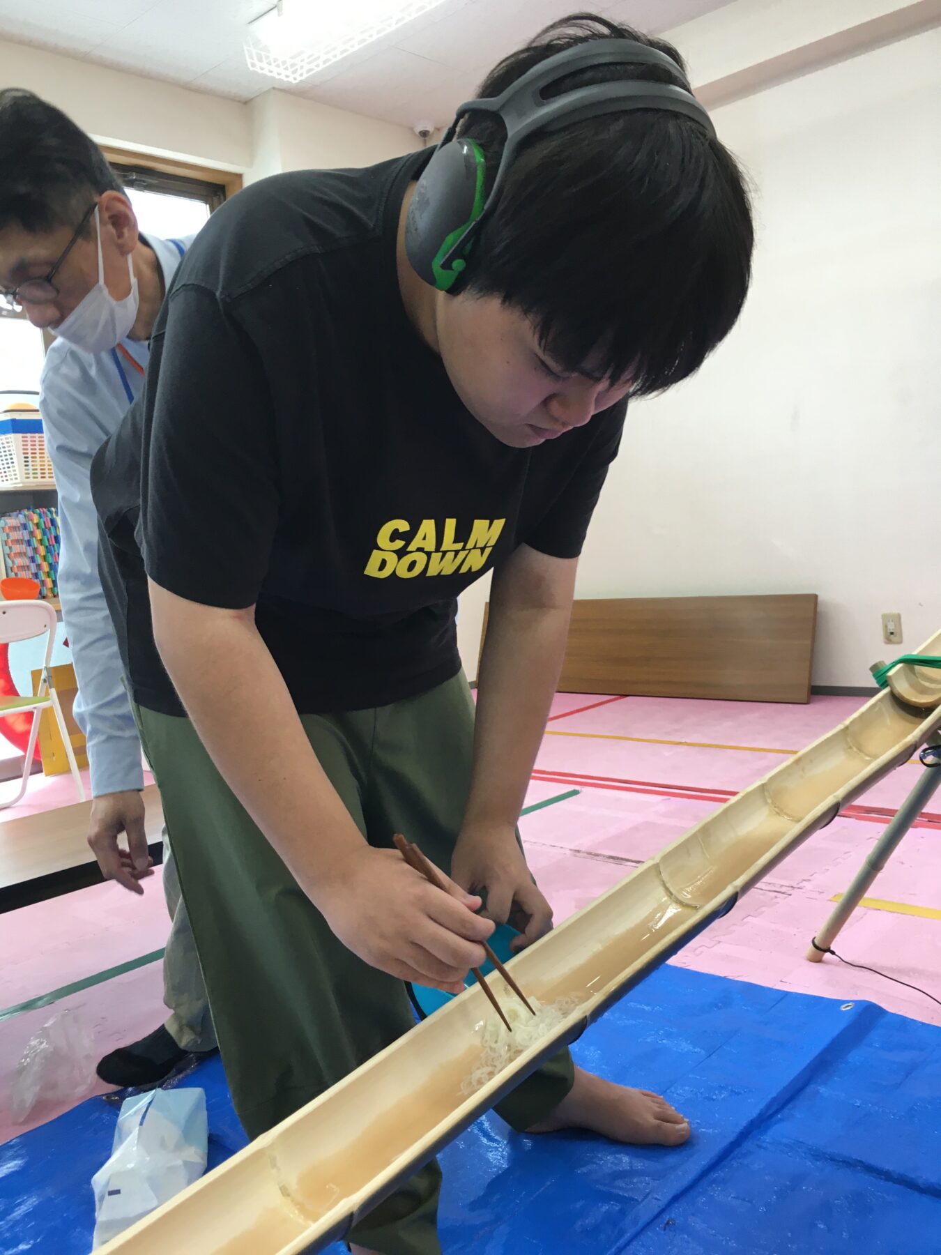 Young person kneeling by a long wooden trough, wearing black shirt reading 'CALM DOWN' and green headphones, handling sticks in a light liquid as another adult observes in a classroom setting.