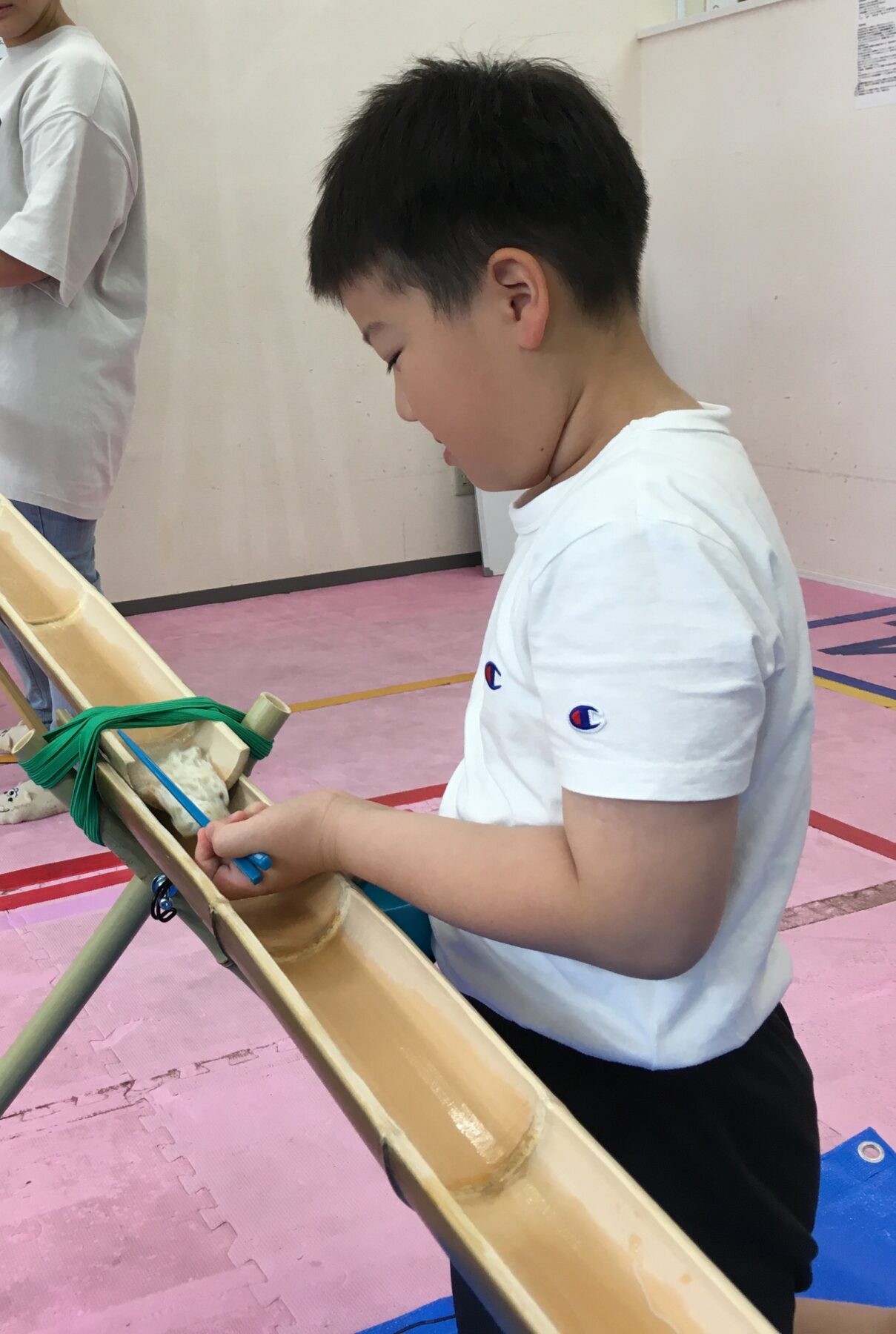 Young boy in a white shirt works on a bamboo craft, tying green straps with a blue tool at a workshop table.