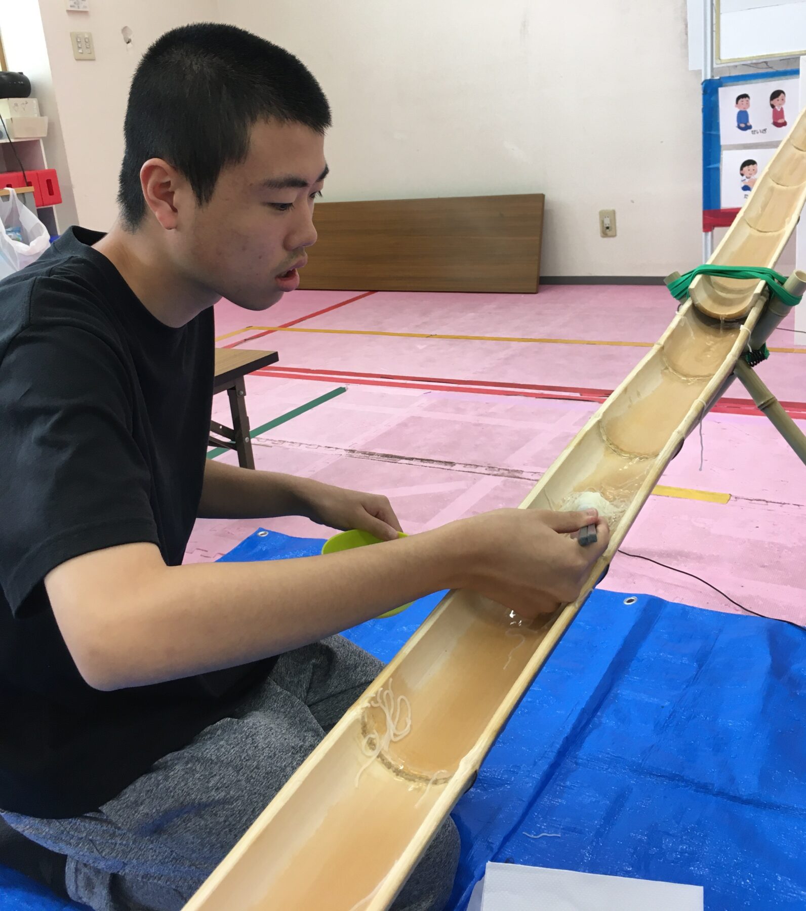 Young man in a black shirt sits at a table, smoothing a long wooden slide/ramp with water channels inside it, indoors on a pink gym floor.