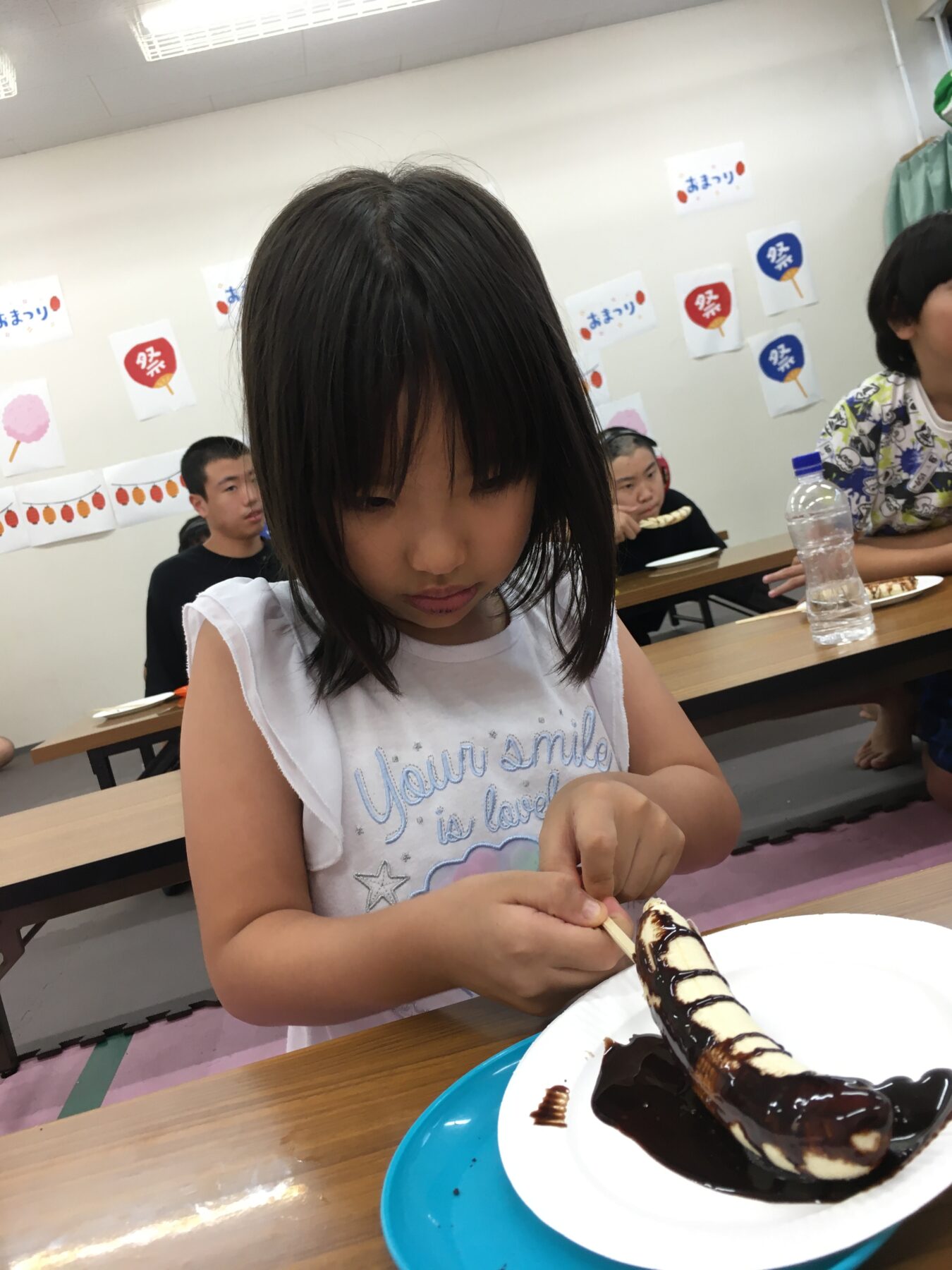 Young girl concentrates on decorating a chocolate-covered banana on a plate at a classroom party with festive posters on the wall in the background.