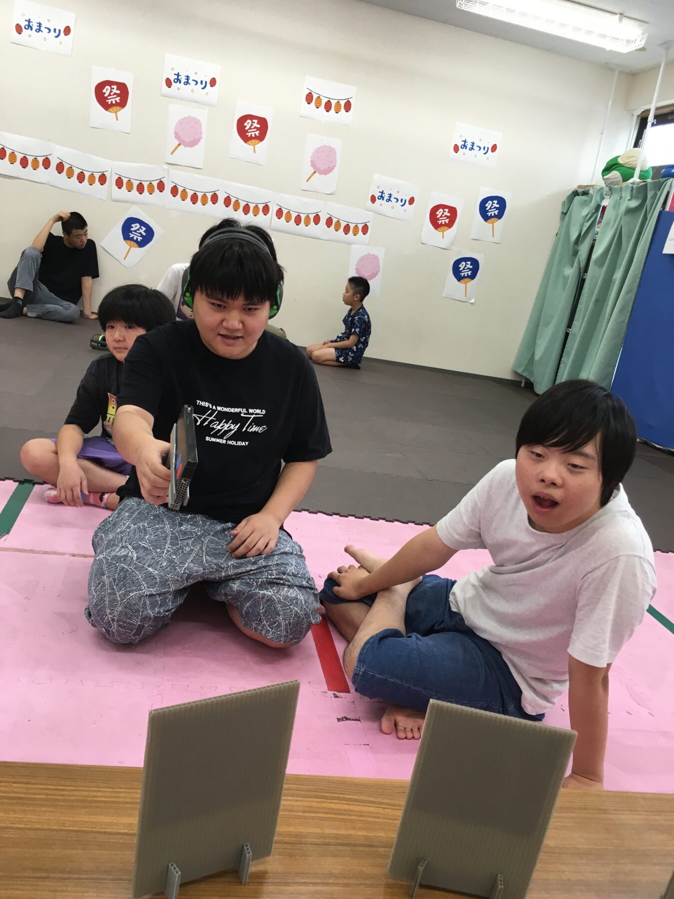 Group of kids and a man with headphones sitting on a pink mat, playing a tabletop game with vertical cardboard panels in a festival-decorated room.