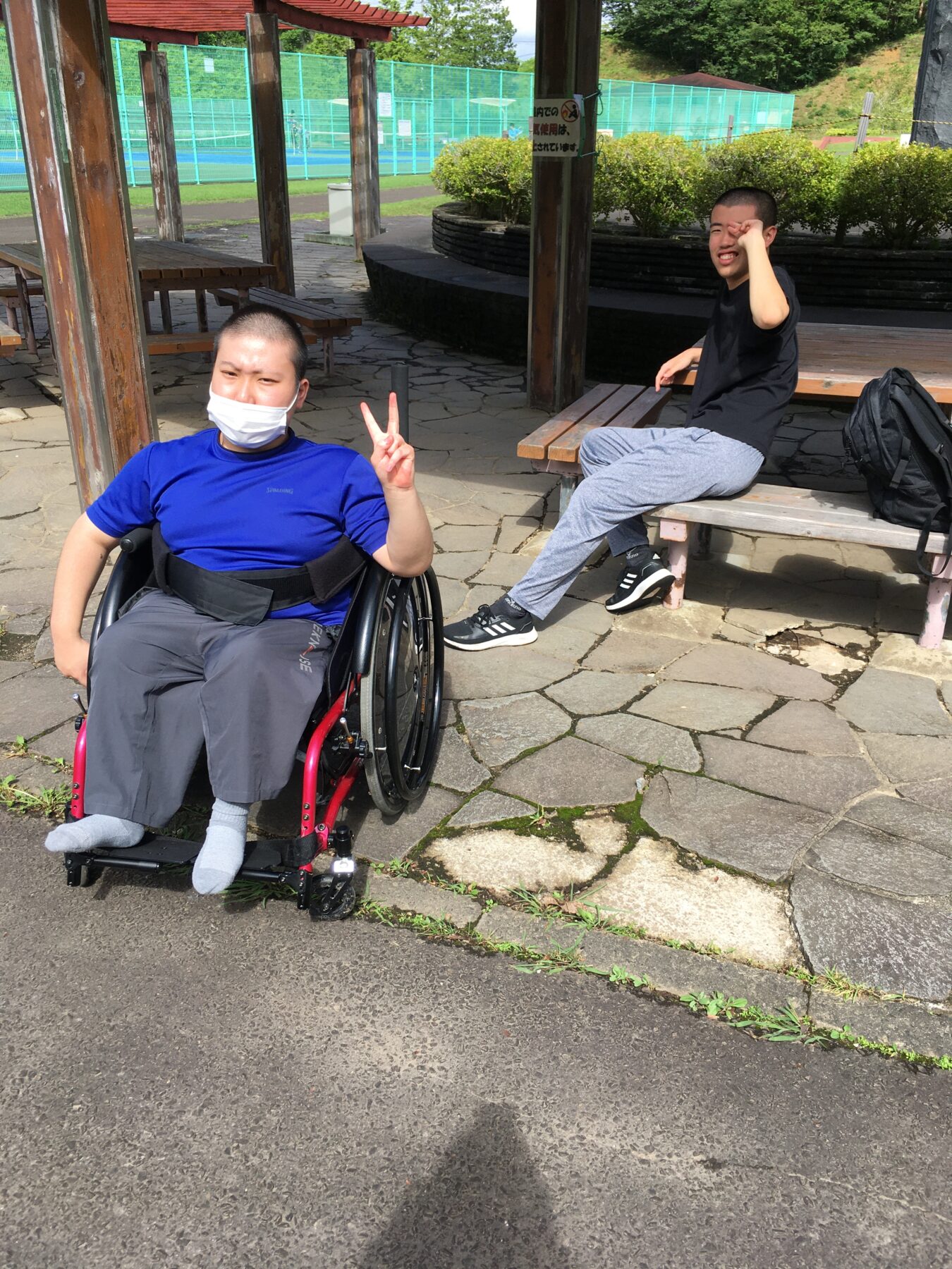 Two young men outside in a park: one in a wheelchair wearing a blue shirt and white face mask making a peace sign, the other sitting on a bench in a black t-shirt and gray sweatpants, shielding his eyes from the sun; tennis courts visible in the background.