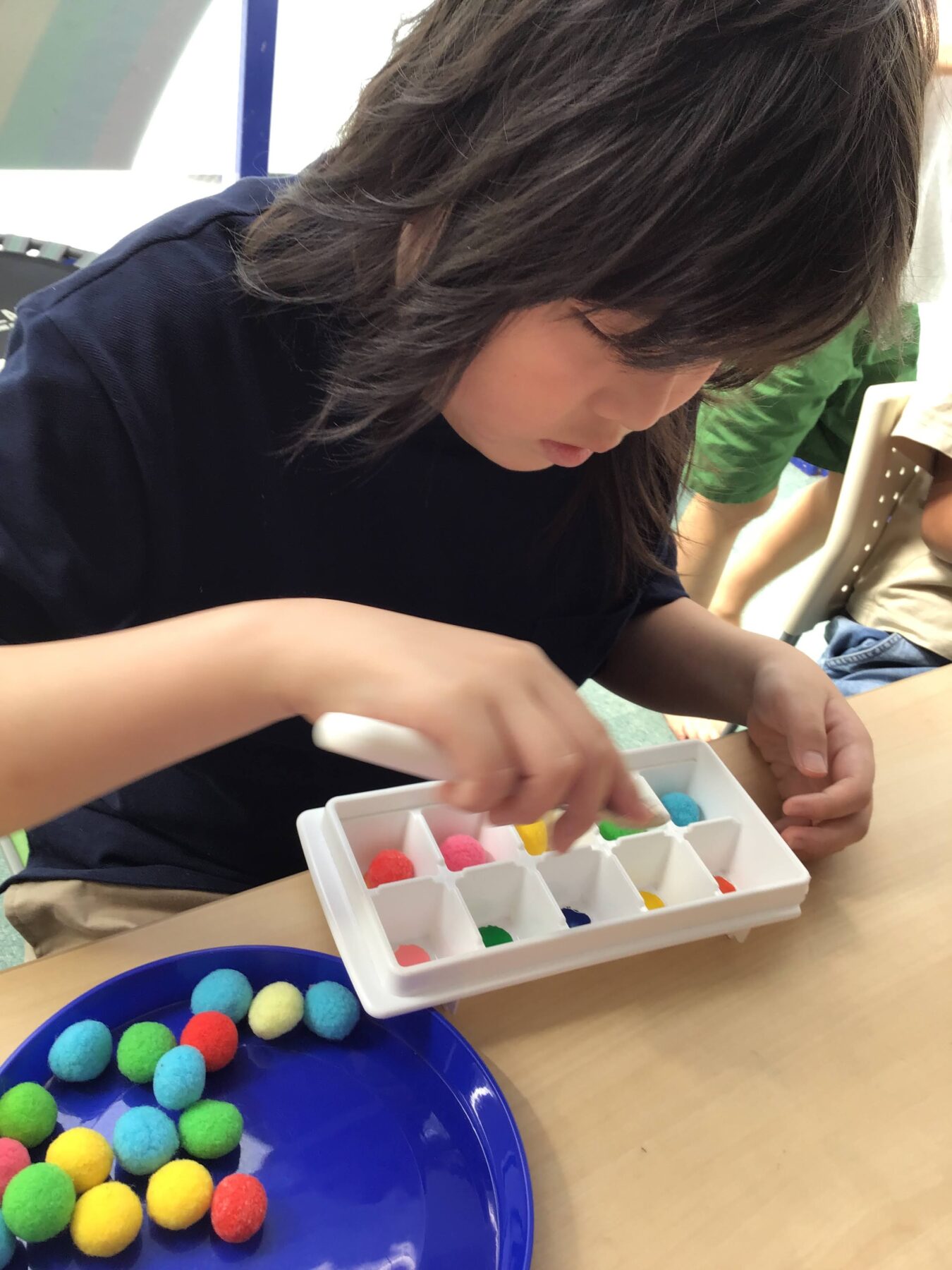 Child's hands sorting colorful pom-poms in a white divided tray during a craft activity, with a blue plate of pom-poms nearby.