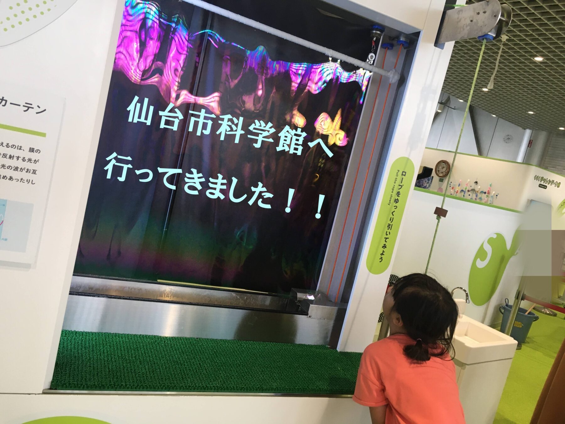 Child in a pink shirt looking at a large colorful screen with Japanese text at a science museum exhibit, green carpet underfoot.