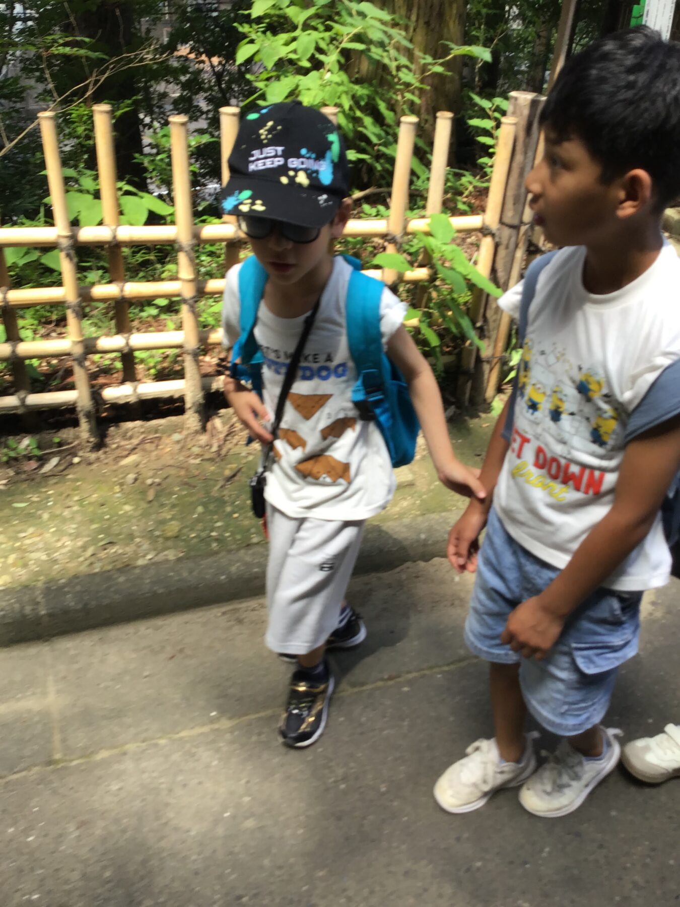 Two kids walk side by side on a sidewalk beside a bamboo fence; one wears a black cap with paint splatters and a blue backpack, the other in a white T‑shirt.