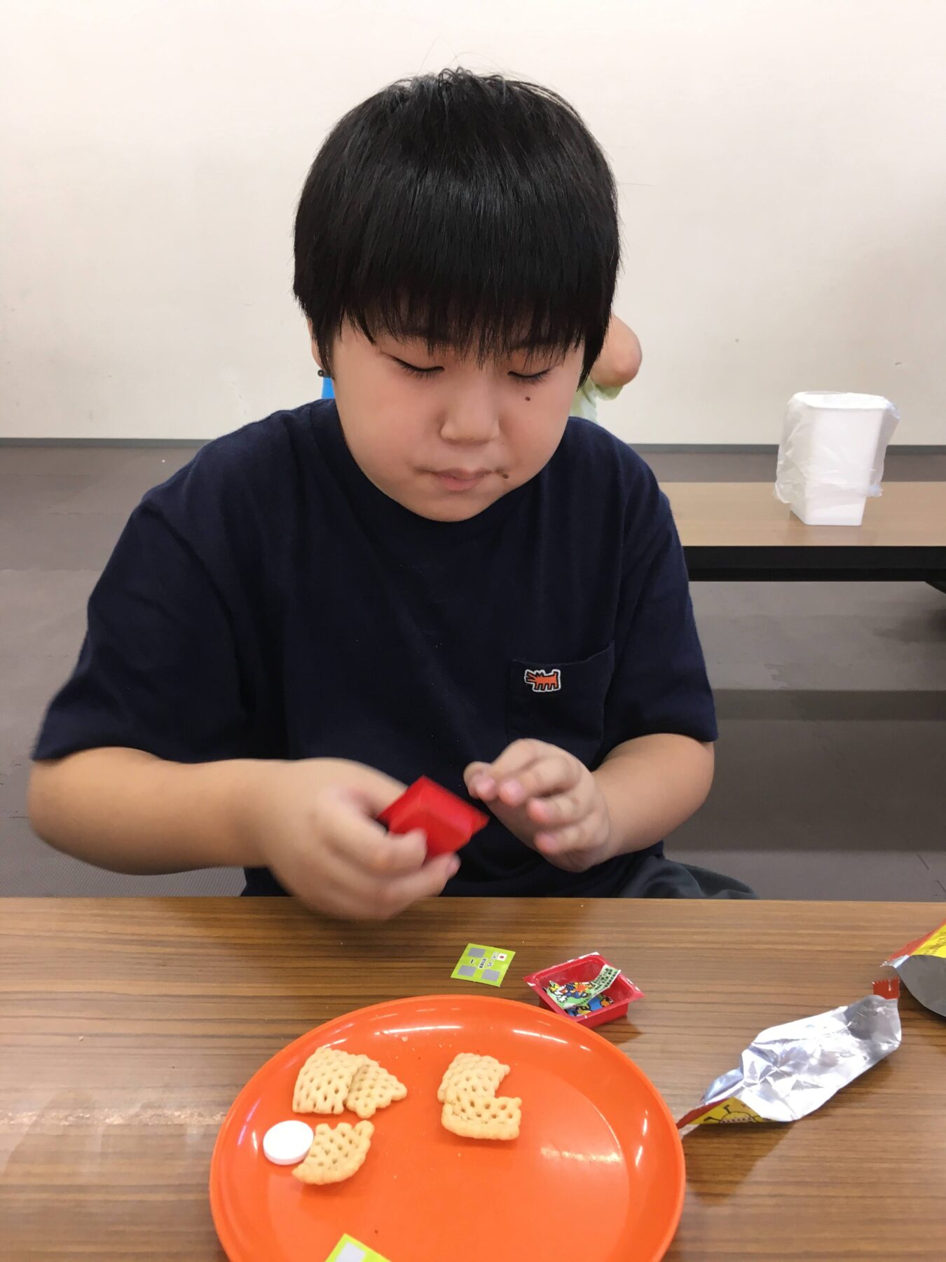 Young child with short black hair and a navy shirt sits at a table, concentrating on a red wrapper in his hands. In front is an orange plate with small square crackers; a few wrappers are on the table.