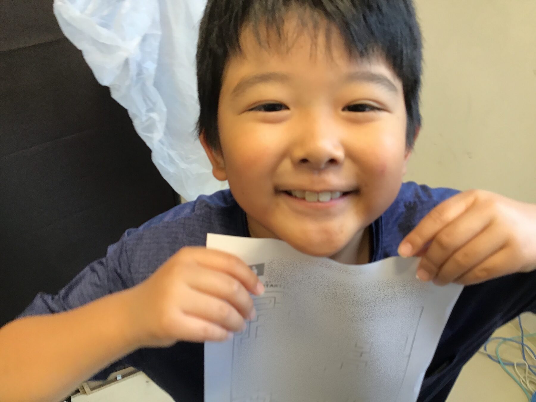 Smiling boy with short black hair holds and tears a gray sheet of paper toward the camera.
