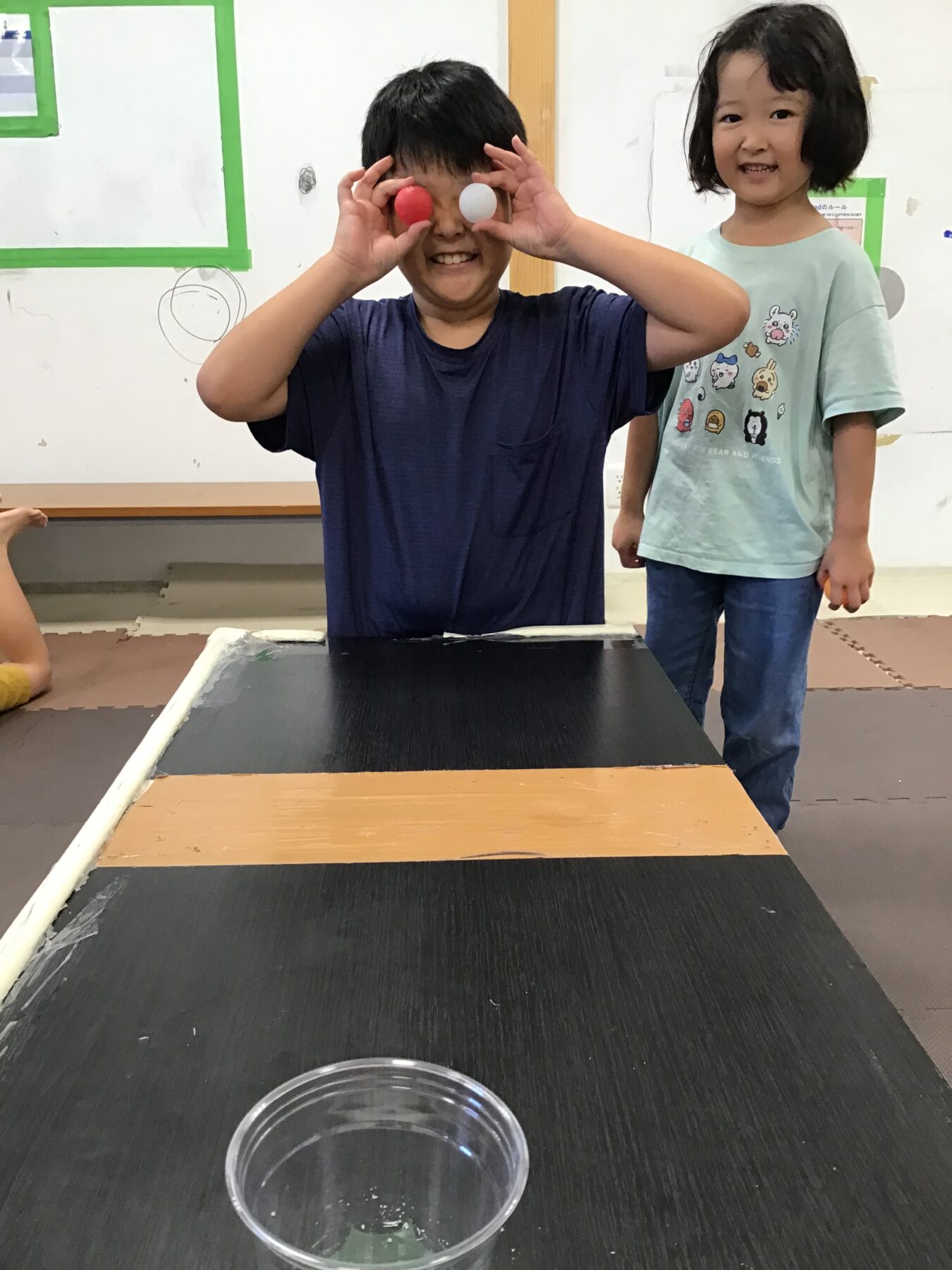 Two kids stand at a black game table; boy in a navy shirt holds red and white balls to his eyes, smiling, while girl in a pastel shirt watches.