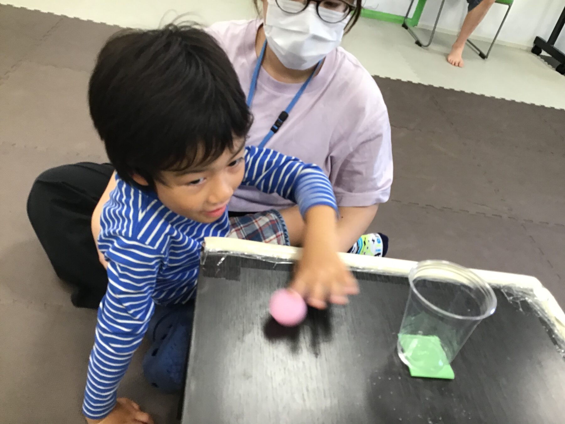 Child kneels at a dark table, reaching for a pink ball while a woman wearing a mask sits behind him, supervising.
