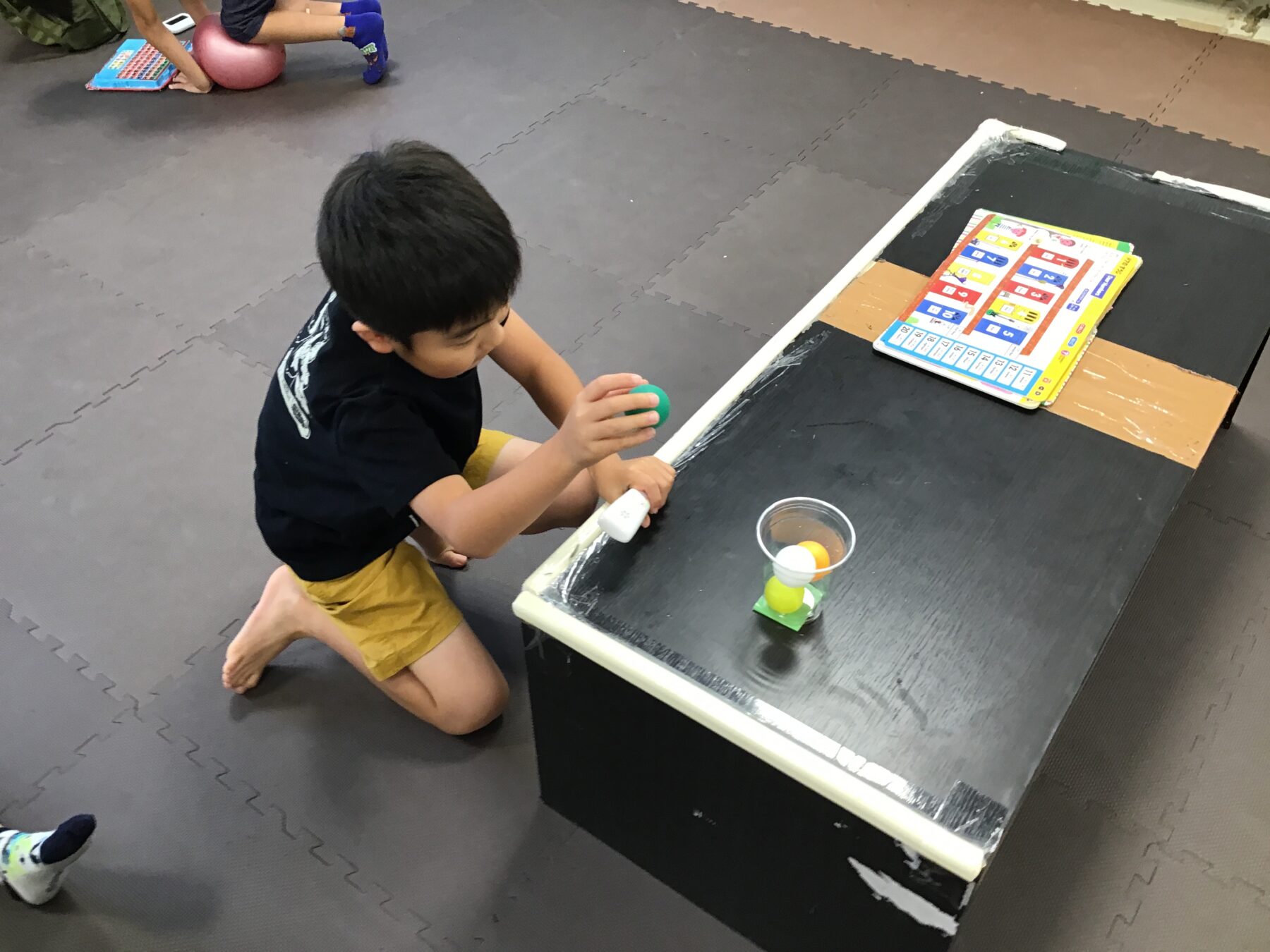 Child kneeling on a padded floor, playing a tabletop ball-stacking game with colored balls and a small ramp table near a colorful activity book on the table.