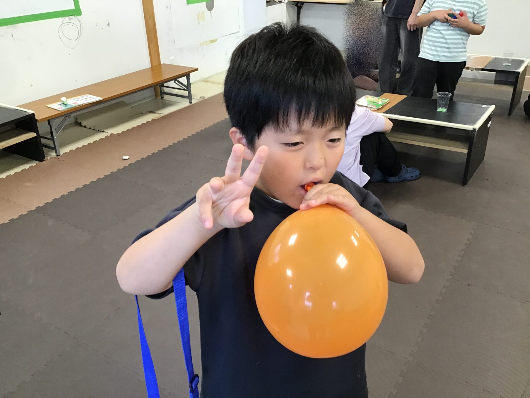 Boy in a dark shirt inflates an orange balloon while making a peace sign with two fingers in a room with foam mats and benches in the background.