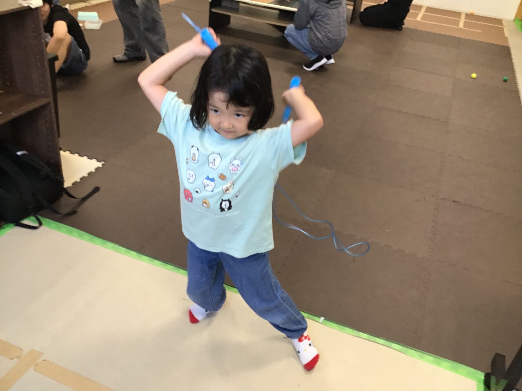 Child wearing a light blue shirt with cartoon faces, blue jeans, and red-toe socks, raising blue ribbons in both hands in a play area with foam mats and other kids in the background.