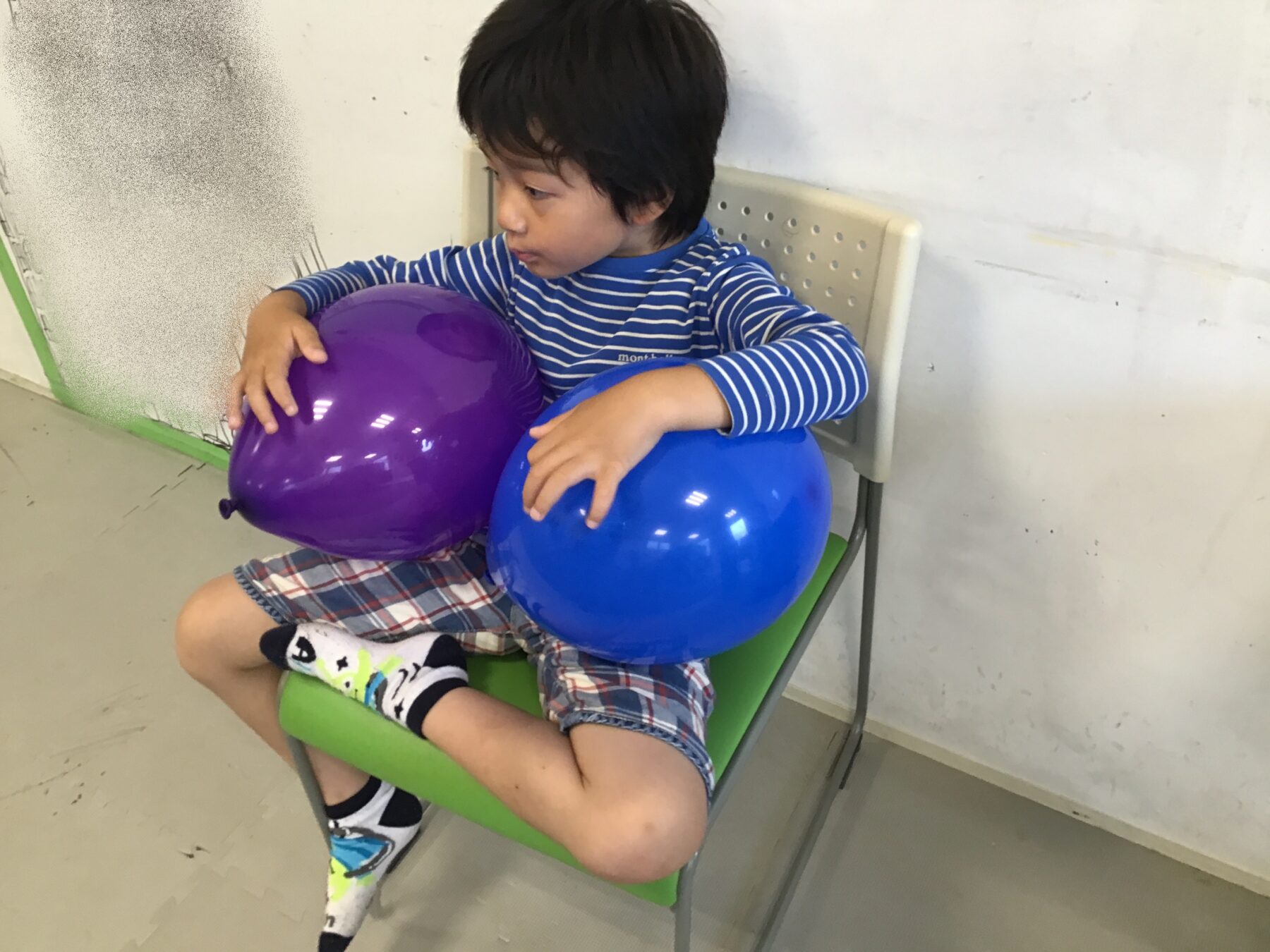 Young boy in a blue striped shirt sits on a green chair, holding a purple balloon and a blue balloon.