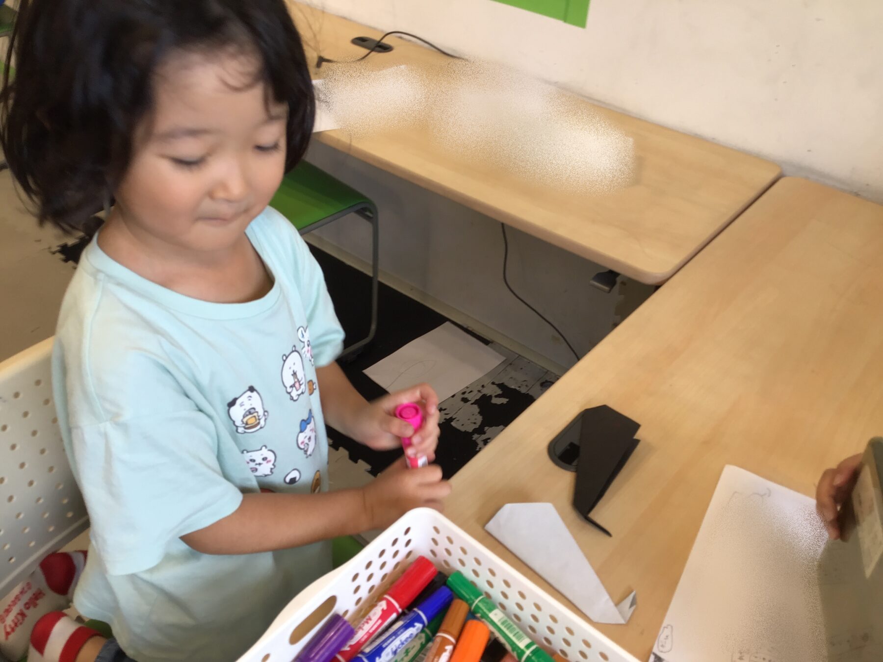 Young girl in a light blue shirt sits at a desk coloring with pink marker, markers in a basket nearby.