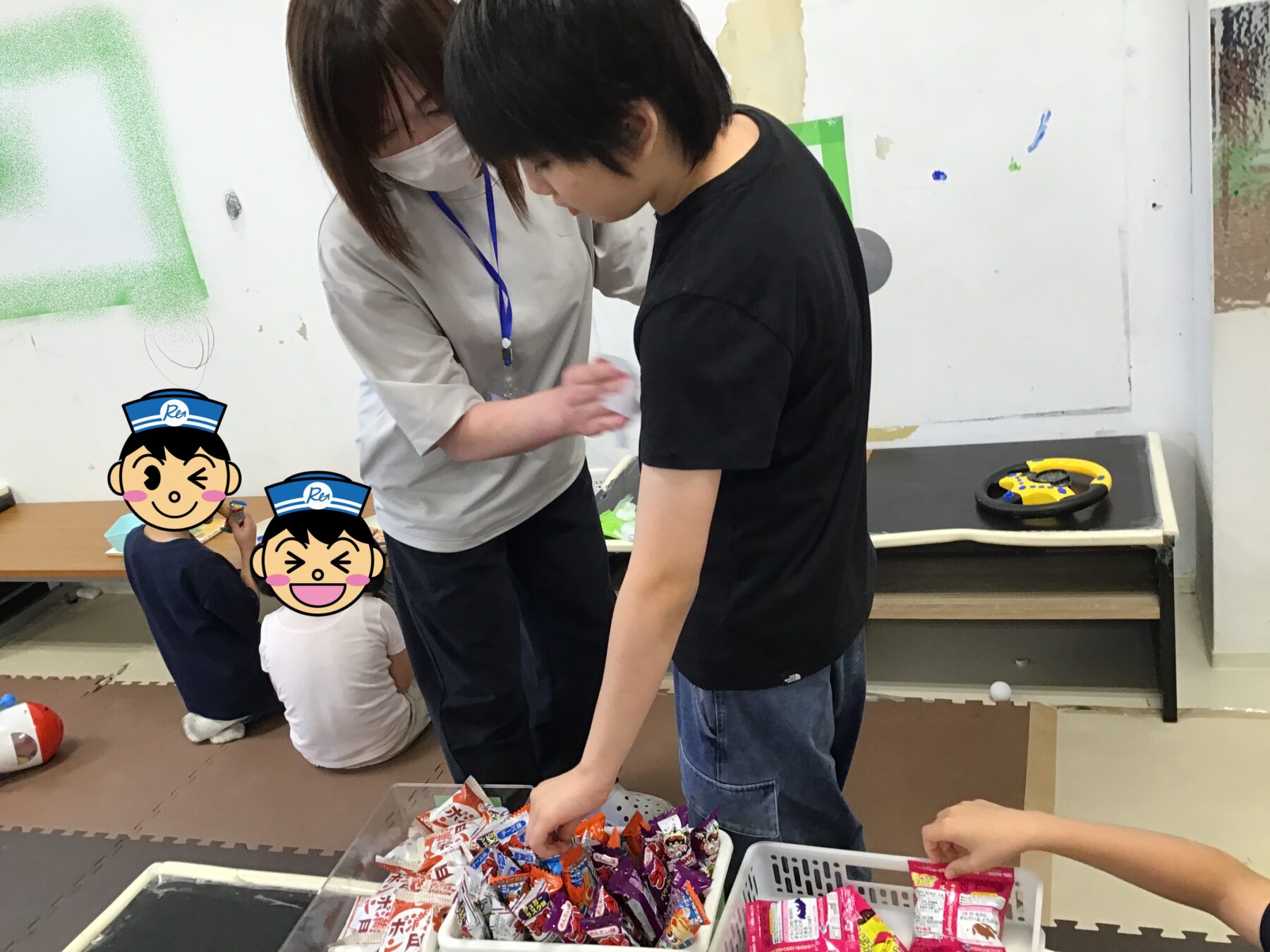 Adults and children gather around a low table sorting colorful candy wrappers during an activity. A steering wheel toy sits on a nearby table in a classroom setting.