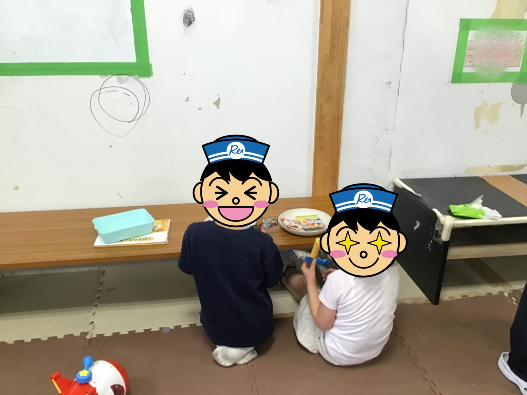 Two children sit on a foam mat at a long wooden table, playing with small toys in a classroom setting.