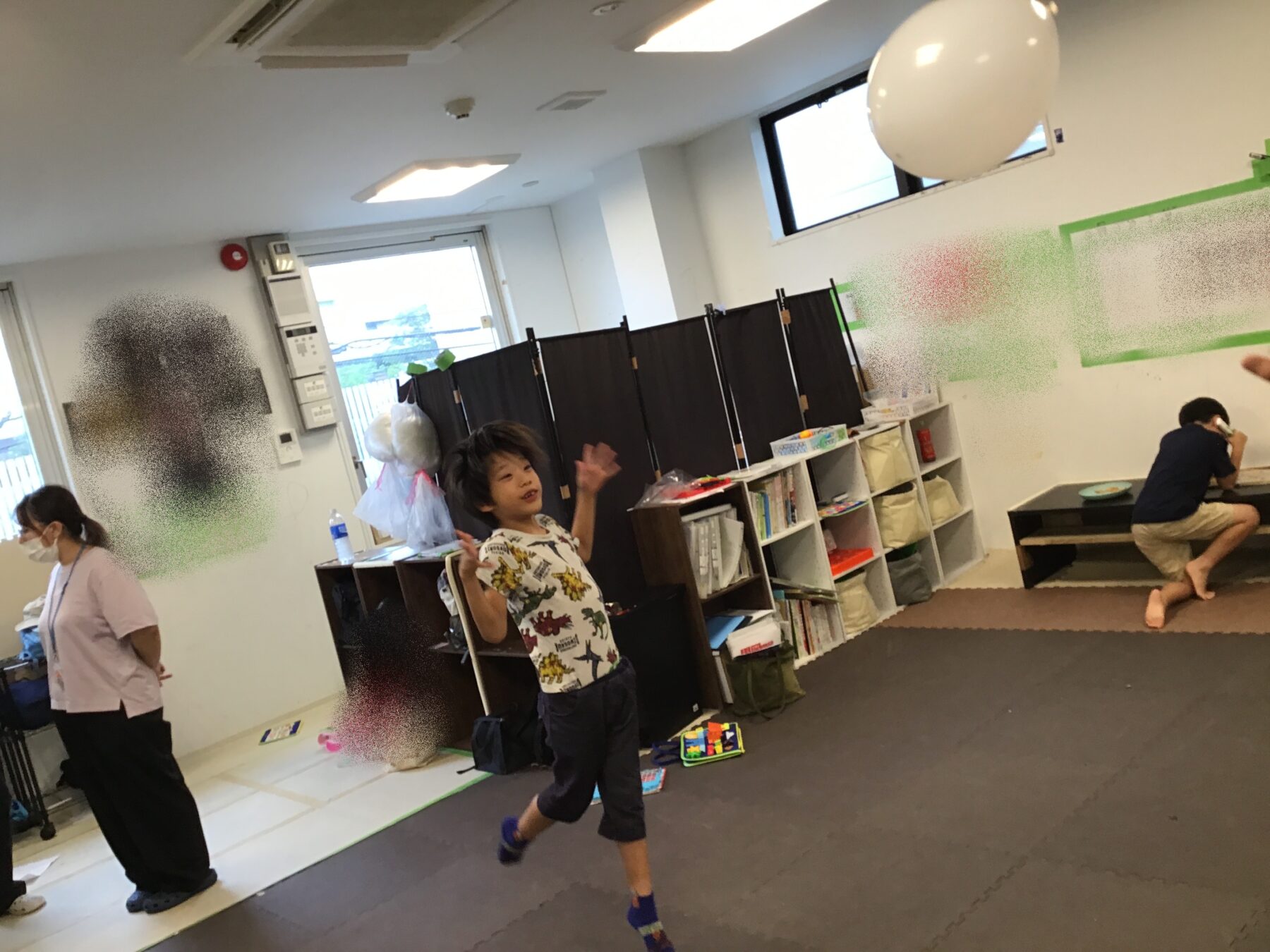 A young boy runs indoors with arms raised in a casual classroom or playroom, other children and adults nearby, shelves with books along the wall, a large white balloon overhead.