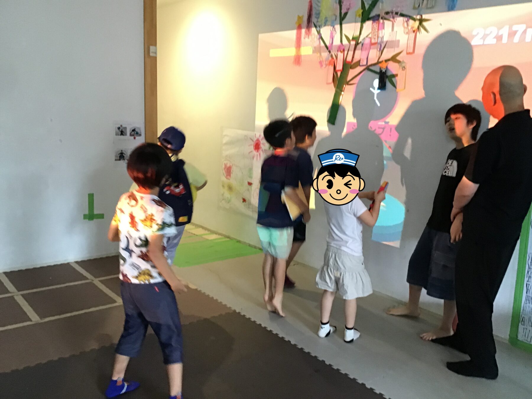 Children and adults gather around a colorful wall art activity in a play area, tracing shadows and placing notes on a tree sculpture.