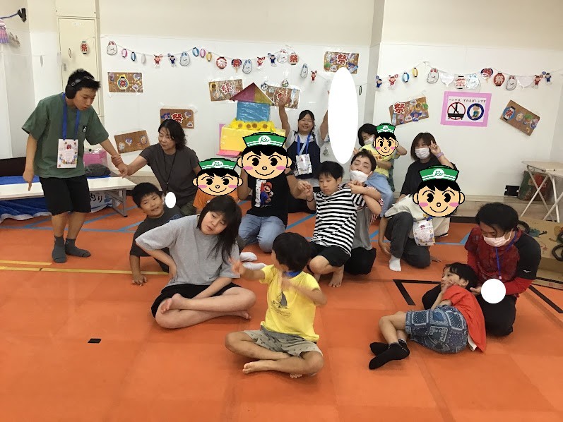 Group of children and adults on an orange mat in a craft-filled classroom, posing for a photo with wall art and a colorful centerpiece in the background.