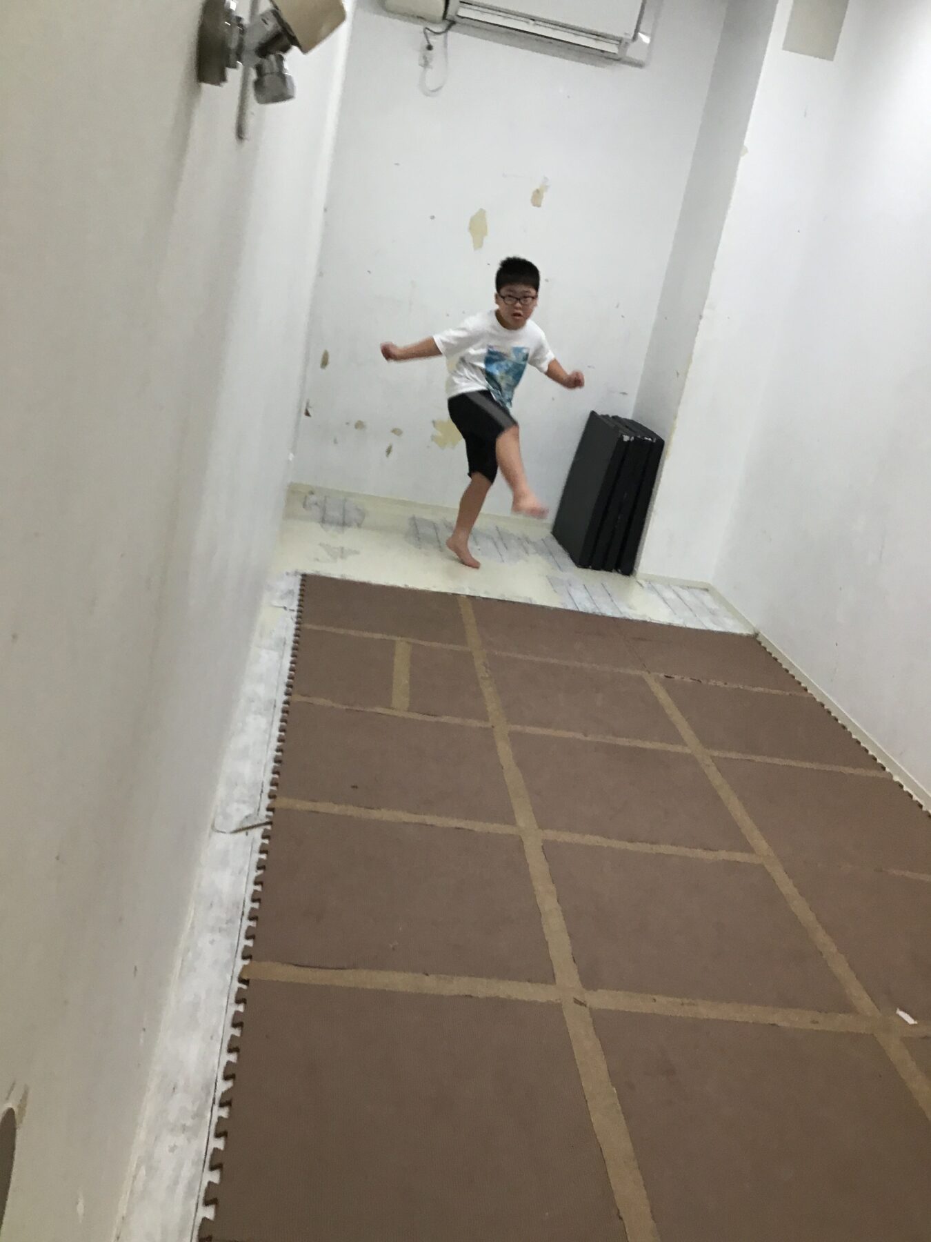 Child in a white shirt and black shorts mid-kick in a narrow hallway with tiled floor mats along the walls.