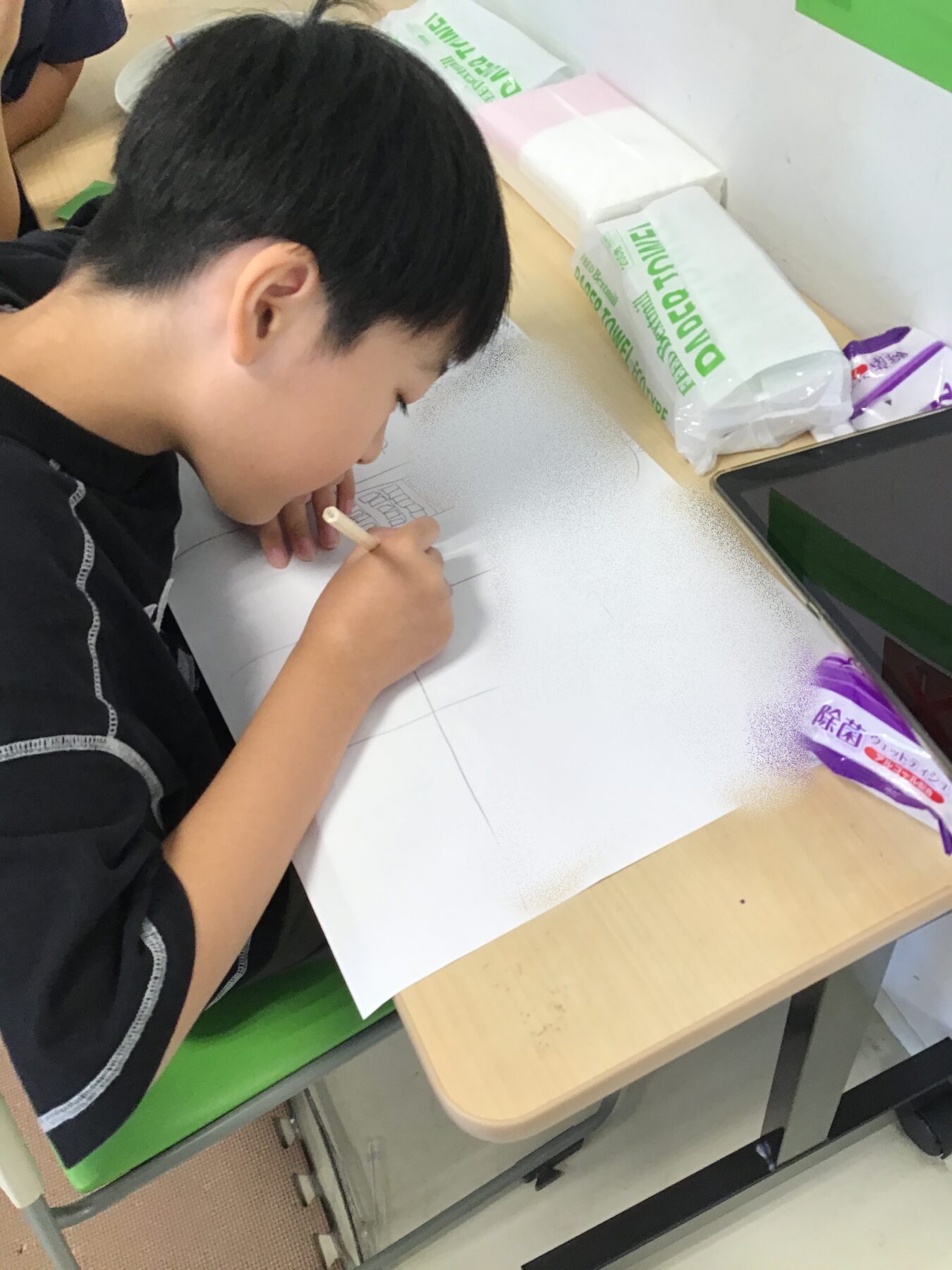 Child leaning over a sheet of paper, drawing with a pencil at a desk with a tablet nearby and boxed supplies on the table?