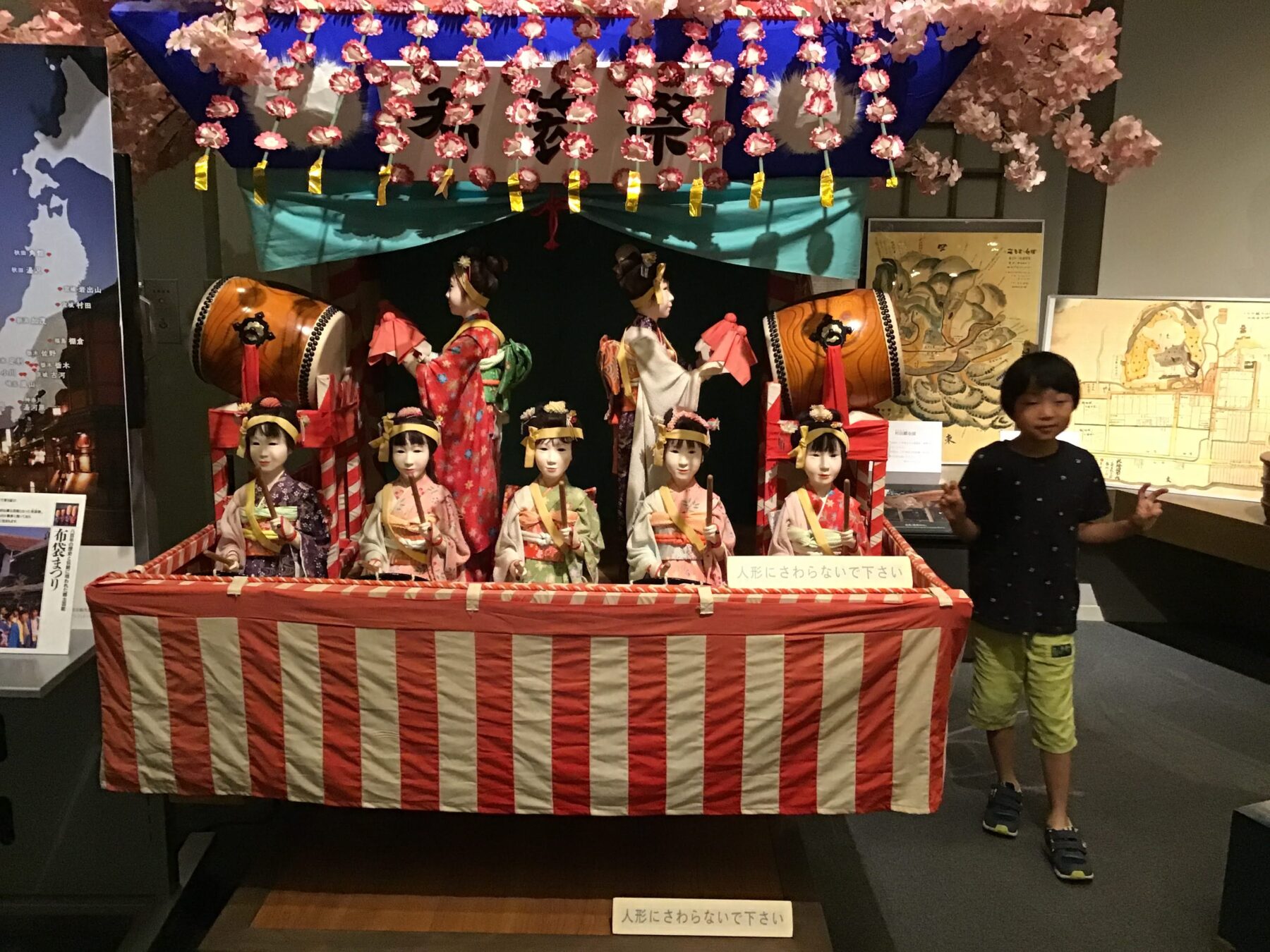 Five traditional Japanese hina dolls in colorful kimonos sit behind a red-and-white striped barrier, with taiko drums and pink cherry blossoms overhead at an exhibition display.