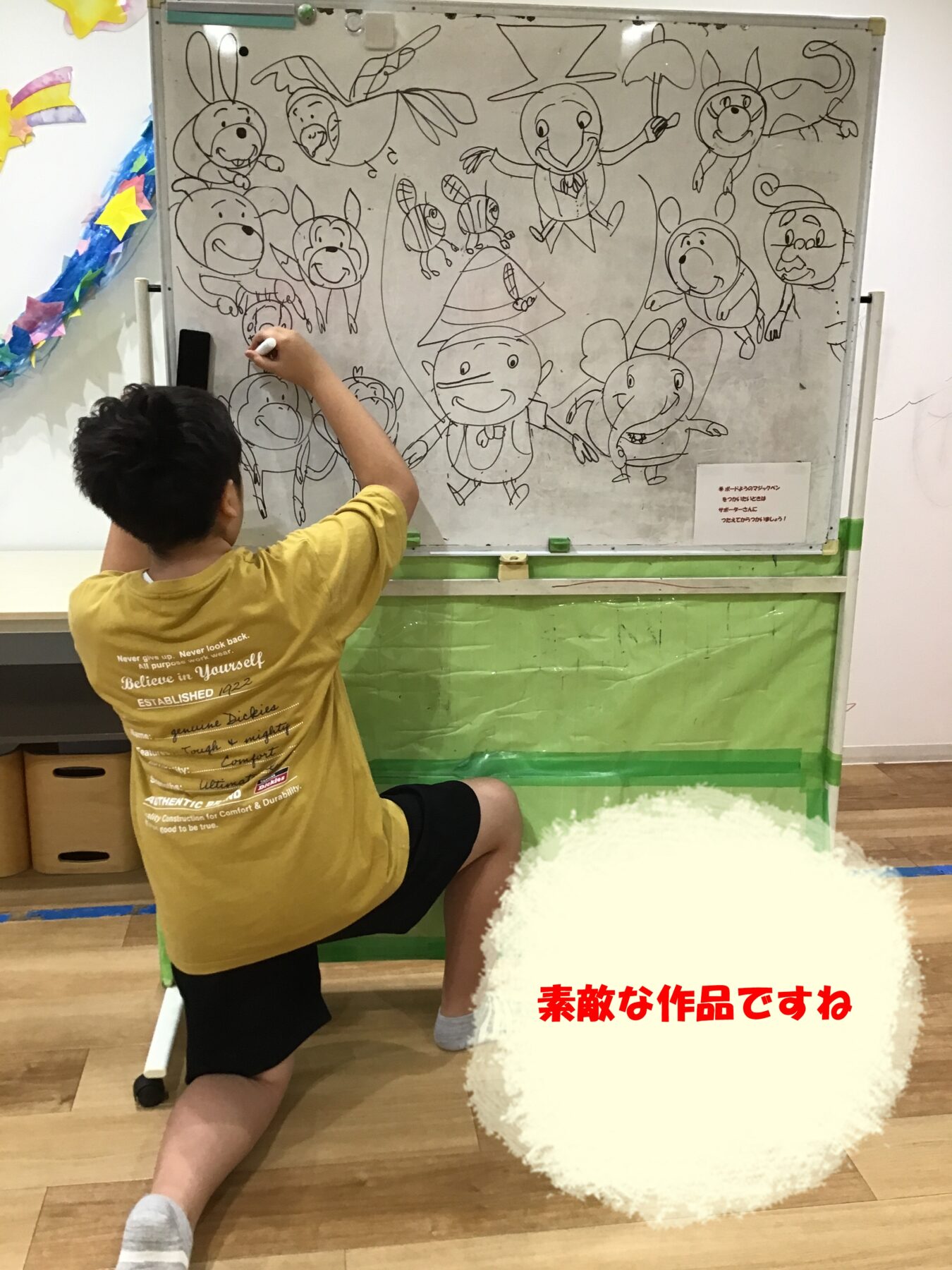 Child in a yellow shirt drawing on a large whiteboard filled with cartoon animals in a classroom setting.