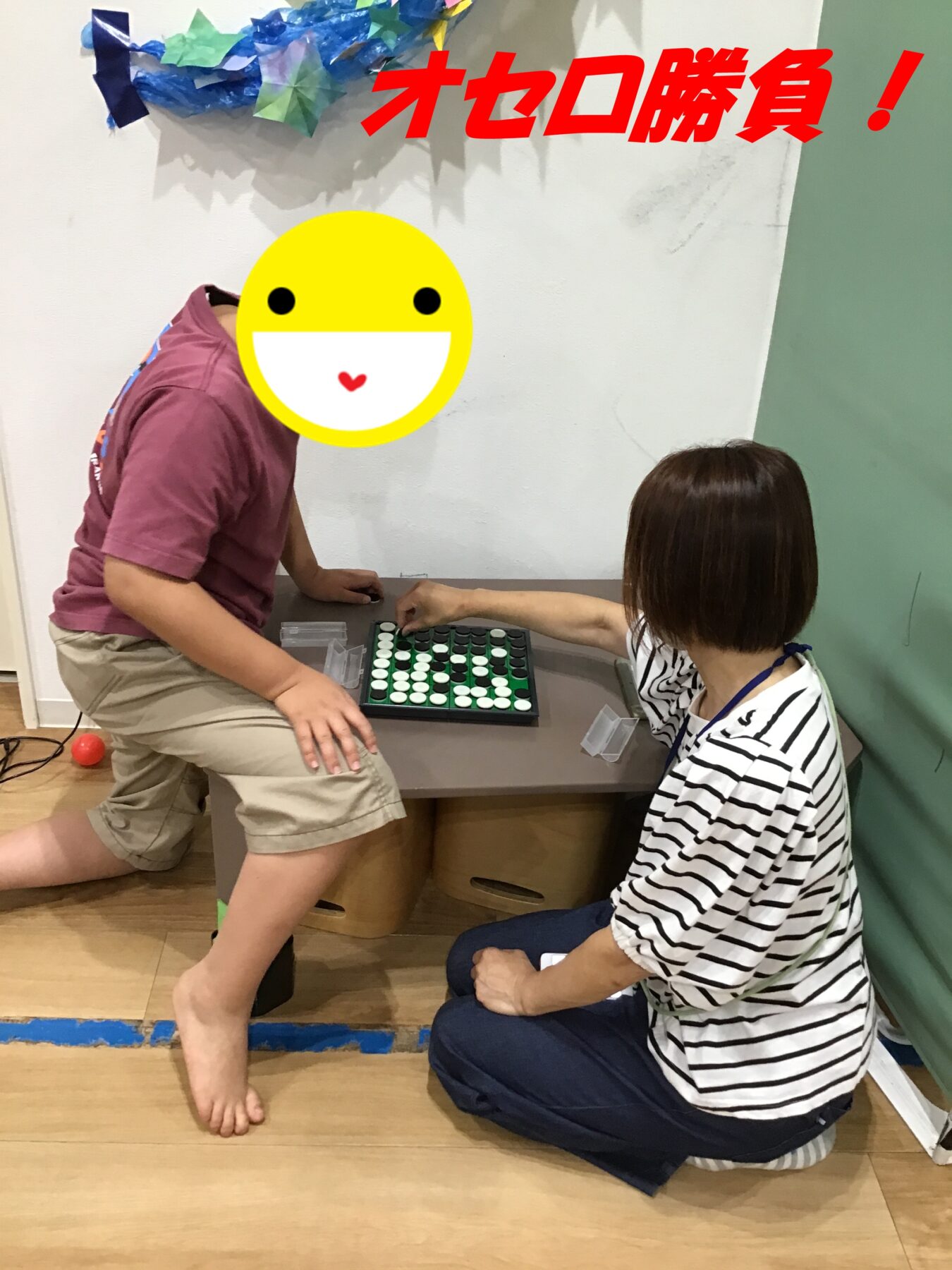 Two people play a board game (Reversi) at a table; one person sits in a striped shirt, the other stands, with a big yellow smiley covering faces in front of decorations on the wall.