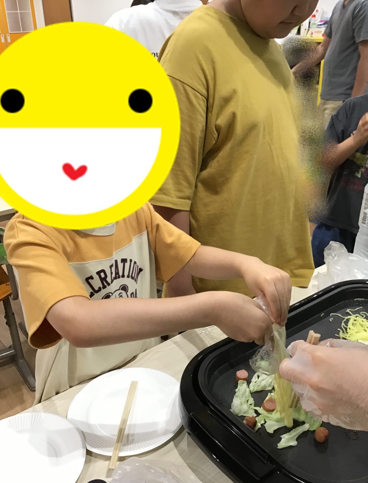 Child in a yellow shirt stands at a table with a large yellow smiley sticker covering their face, helping to prepare food on a griddle with cabbage and small sausages nearby.