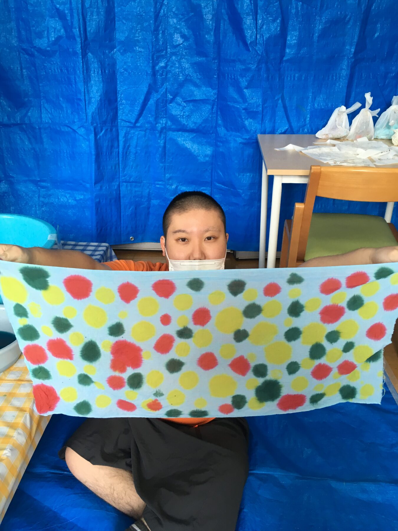 Person with short hair and a mask holds up a bright polka-dotted banner in front of them, sitting in a blue-tarped room.