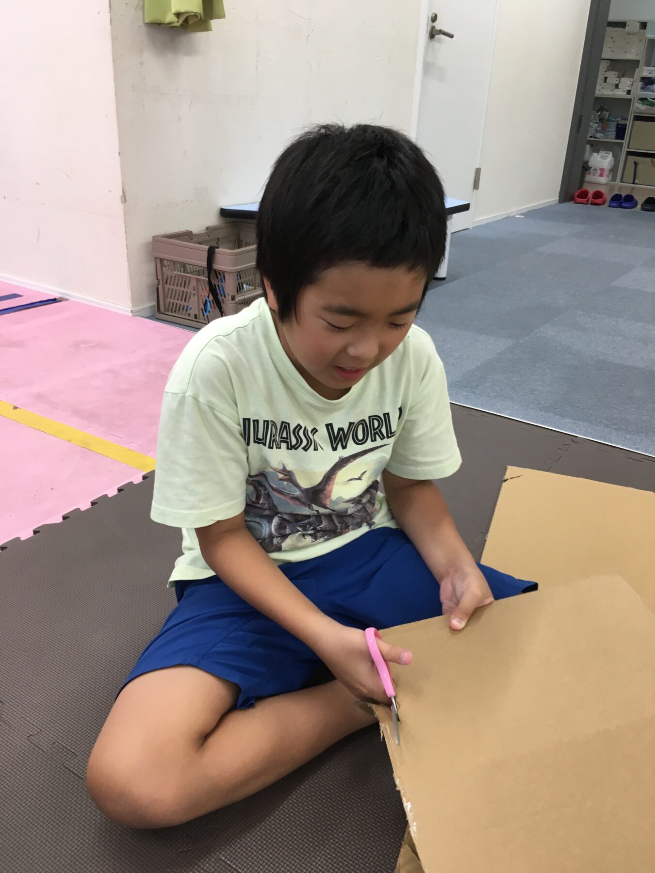 Boy sitting on a foam mat, cutting a large piece of cardboard with pink scissors in a workshop-like room.
