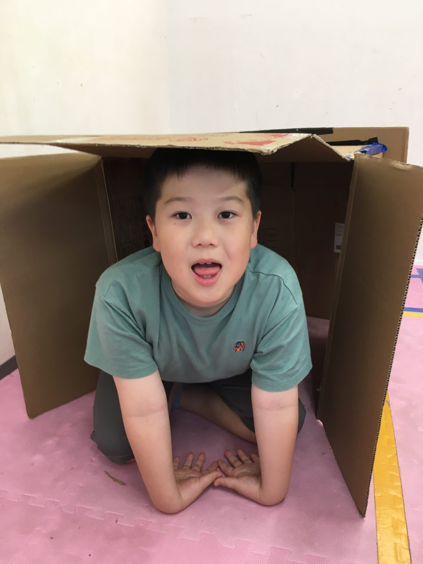 Boy sits inside a cardboard box tunnel, looking at the camera with mouth open in a playful expression on a pink foam mat.