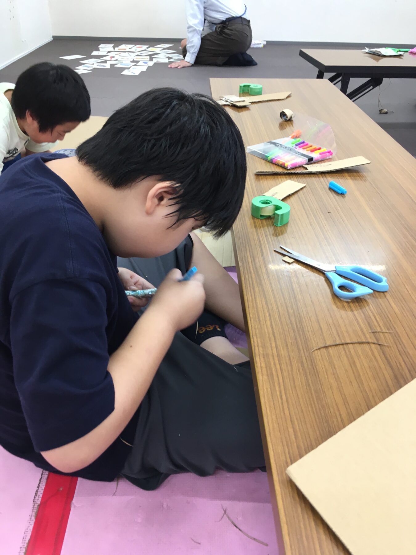 Young boy sits at a craft table, drawing with a blue marker; scissors, tape, and supplies are nearby on the table.