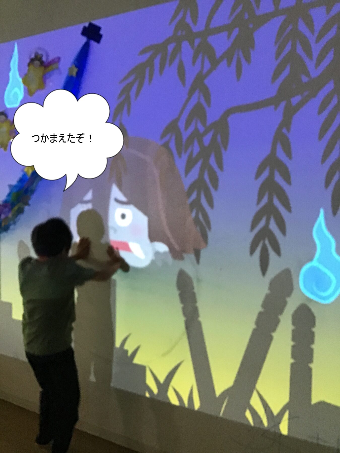 Person standing sideways, reaching toward a large animated wall projection of a girl, with a speech bubble that reads 'つかまえたぞ！'