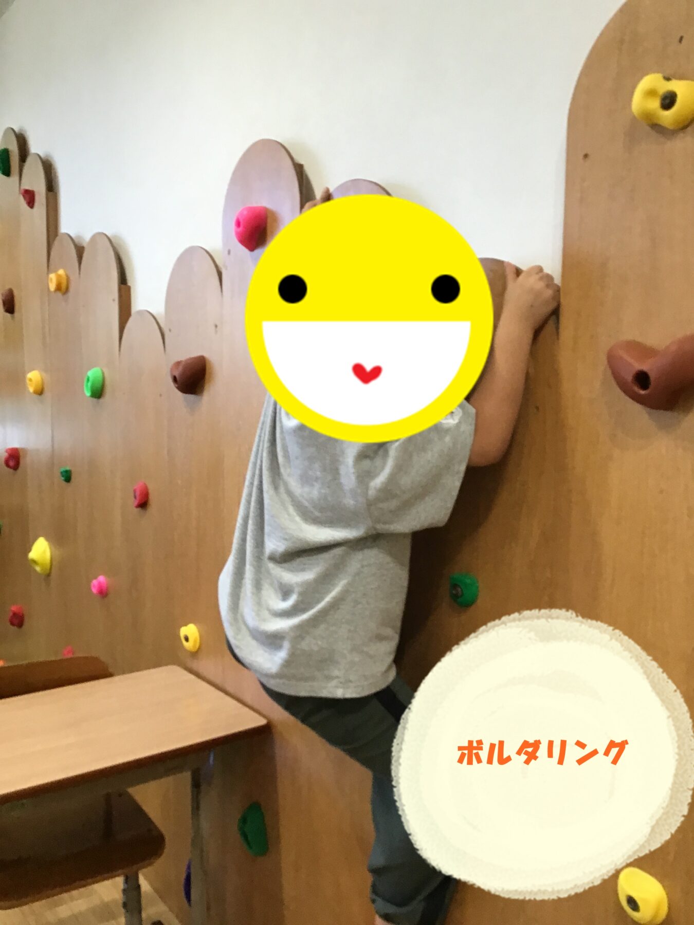 Child wearing a gray shirt climbs a wooden indoor bouldering wall with colorful holds; face is covered by a large yellow emoji sticker.