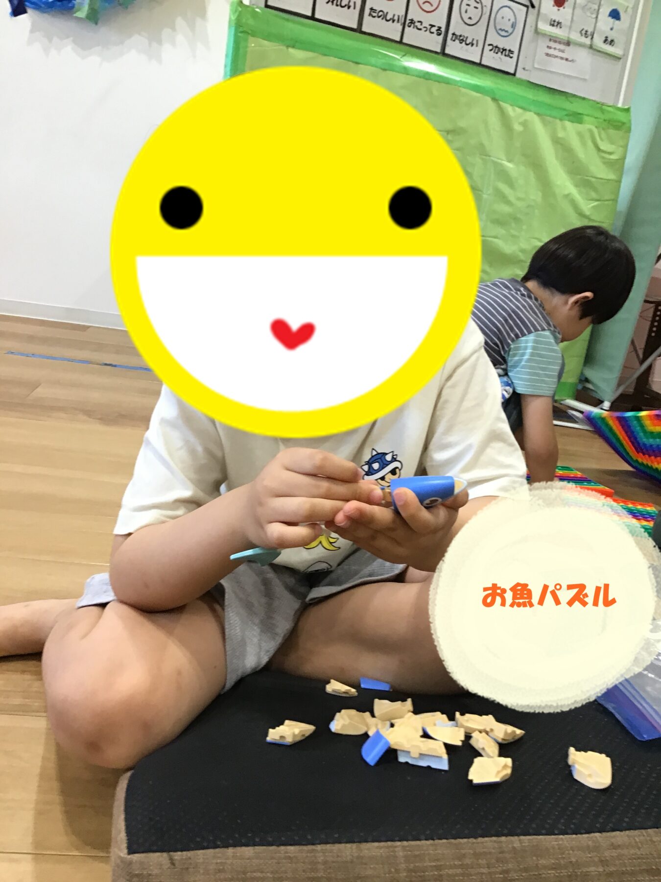 Child sitting cross‑legged on a mat, assembling small wooden puzzle pieces with a large yellow emoji covering their face.