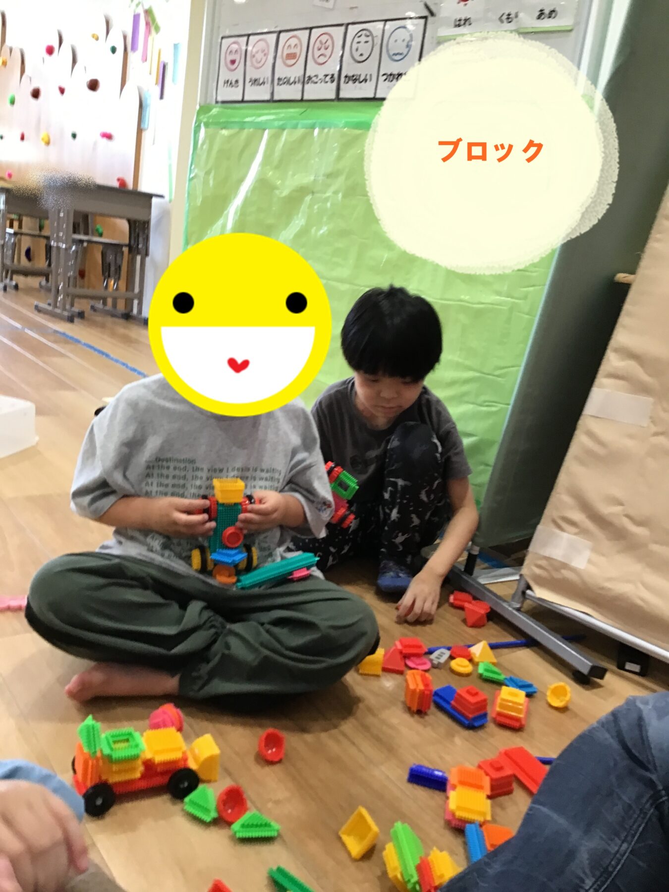 Two kids sit on the floor building with colorful interlocking blocks in a playroom; blocks are scattered around them and a climbing wall is in the background.