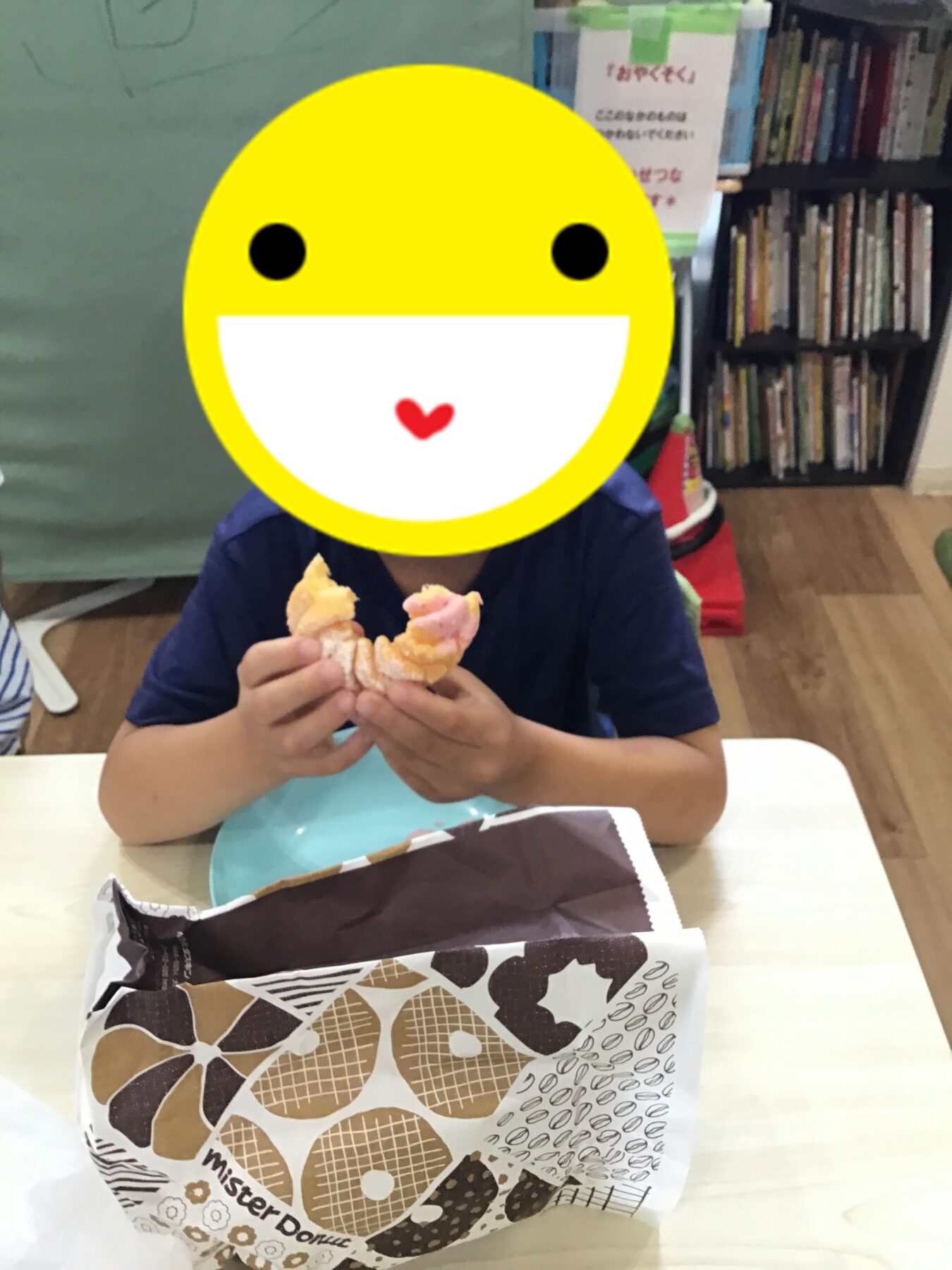 Child seated at a table, eating a donut; a large yellow smiley overlay covers their face, with bookshelves in the background.