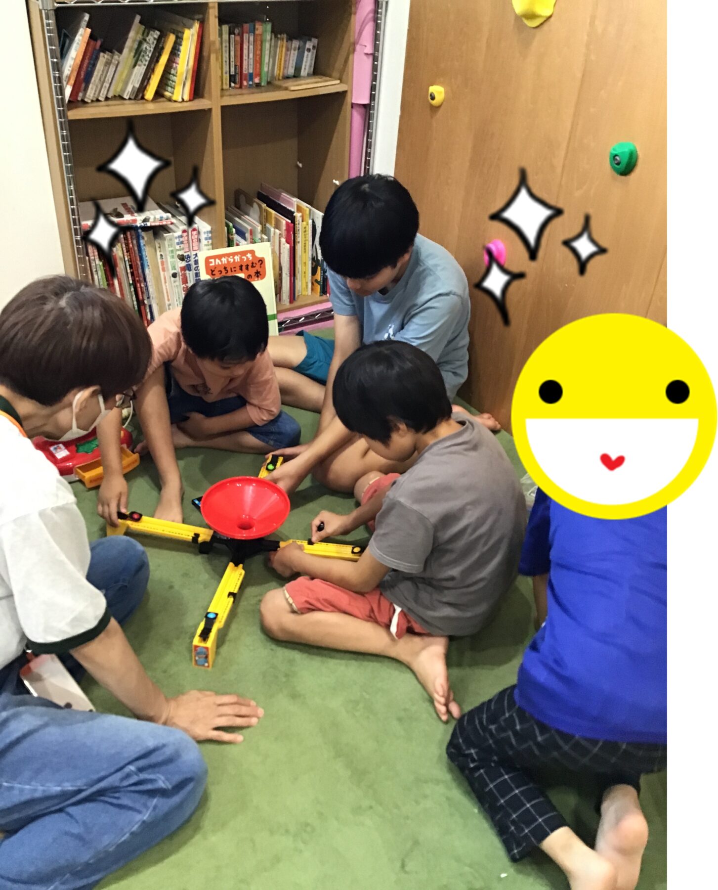 Group of children seated on a green carpet, building a yellow marble-run track with toys while an adult watches in a book-filled room.